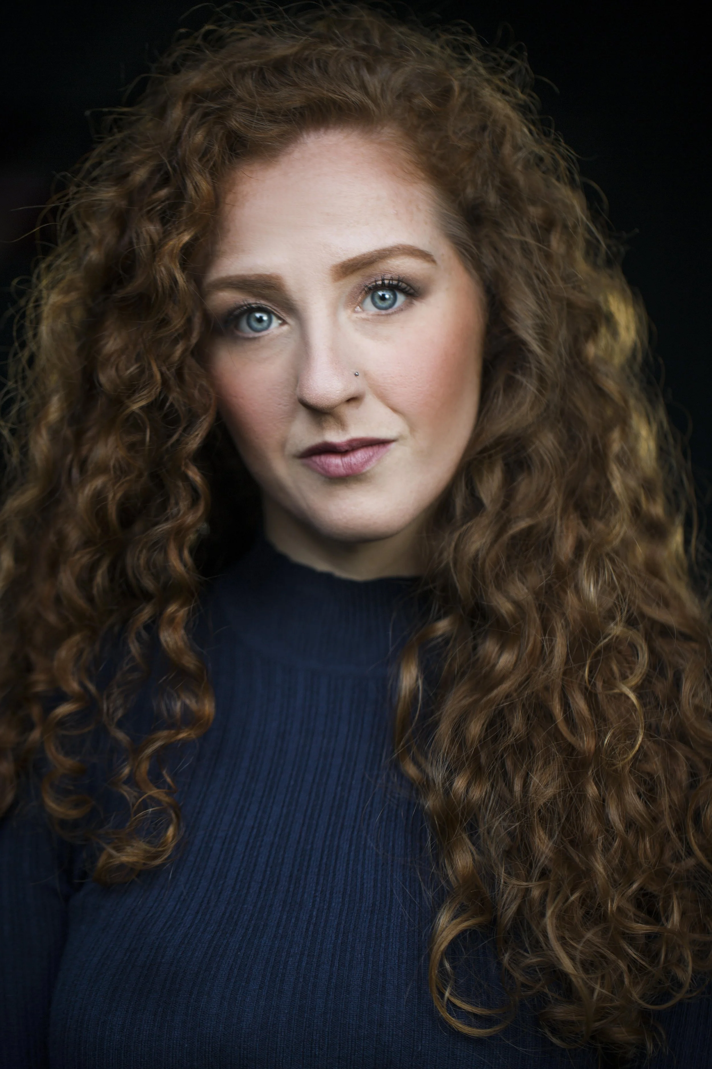 Portrait of a woman with long, curly red hair, blue eyes, wearing a navy blue top, against a dark background.