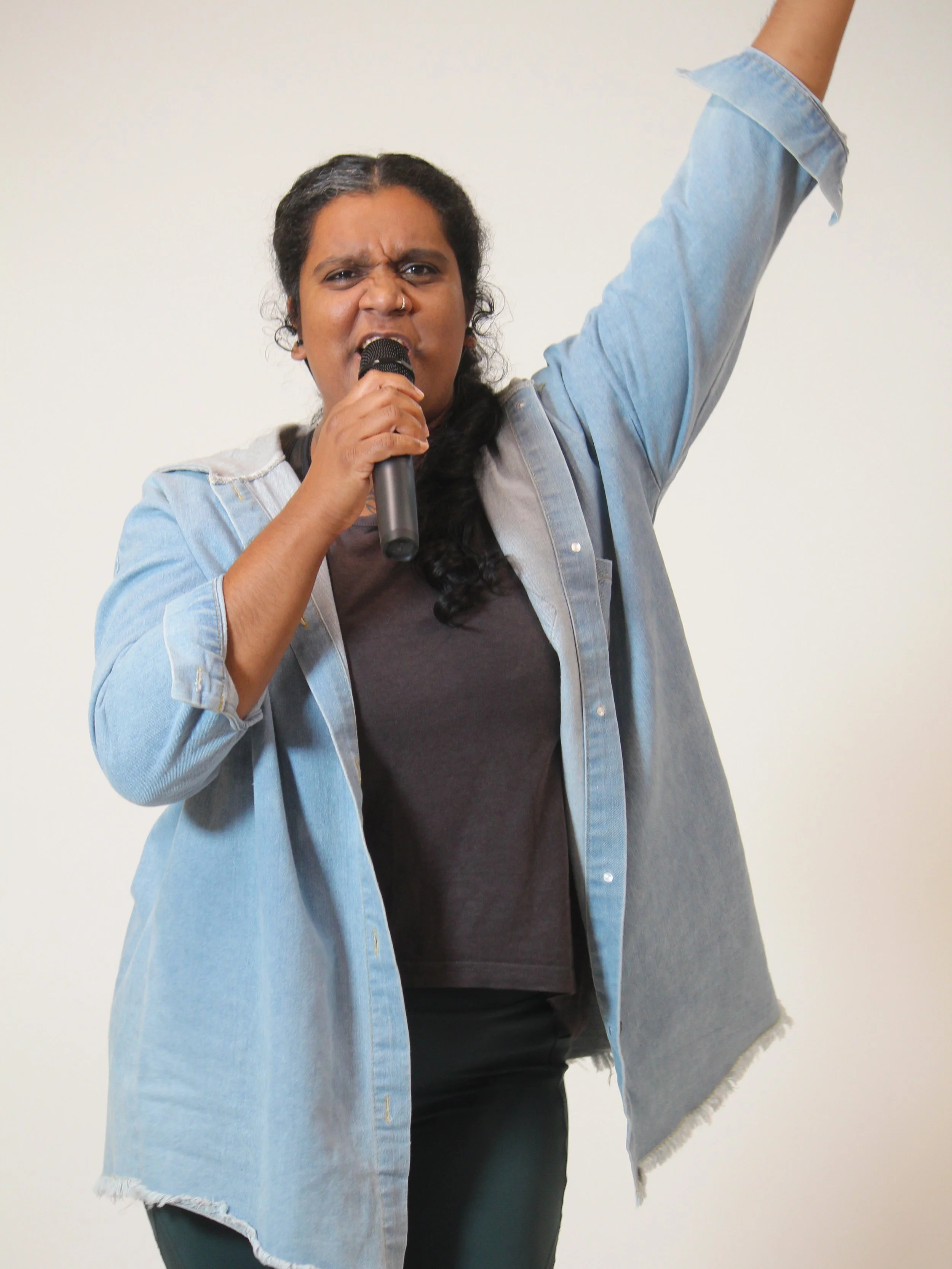 A woman with dark curly hair, wearing a denim jacket over a dark shirt, holds a microphone with one hand and raises her other arm in the air, appearing to sing passionately against a plain white background.