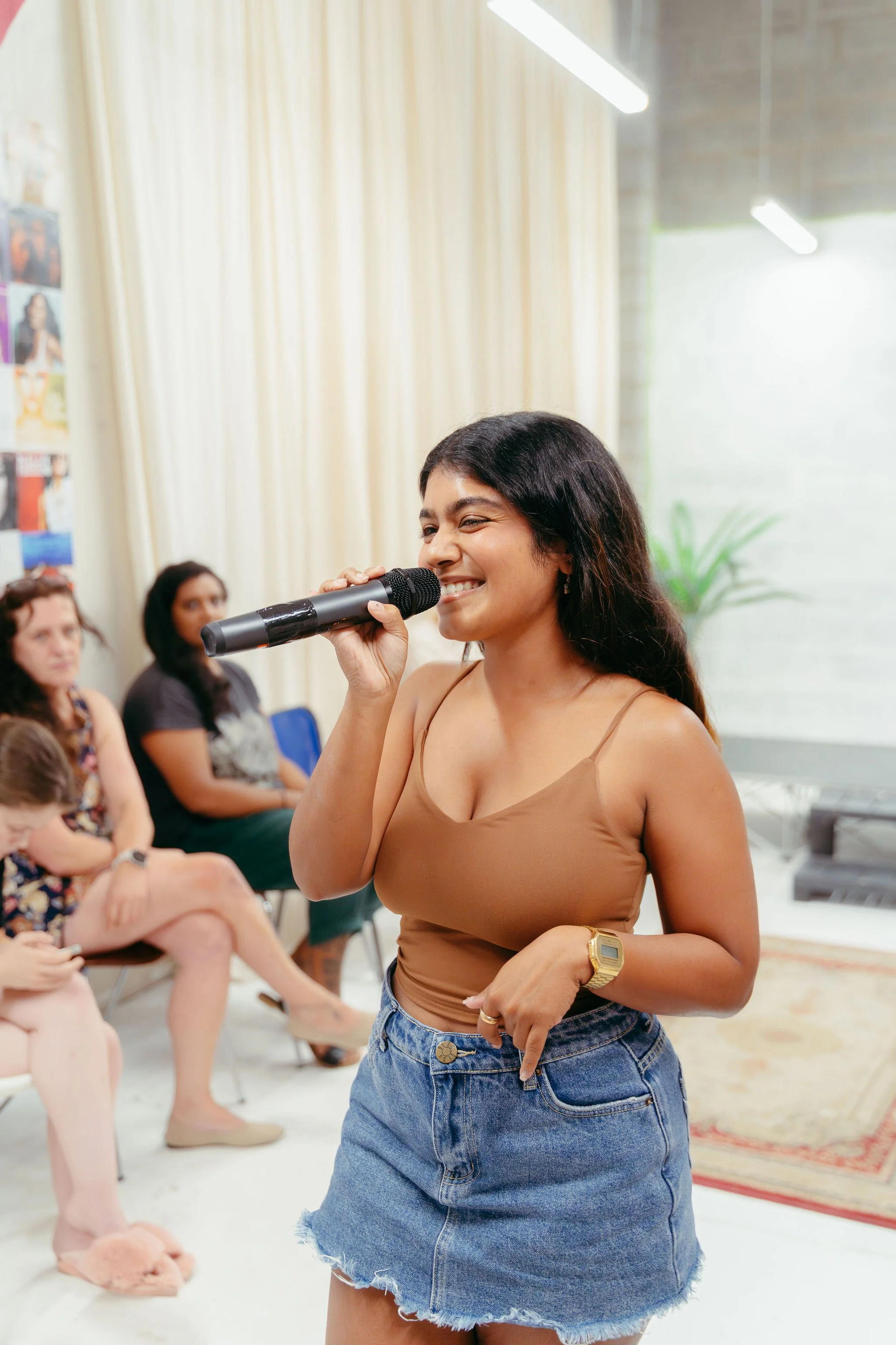 Young woman smiling while holding a microphone during a group event in a bright room with curtains and artwork on the wall.