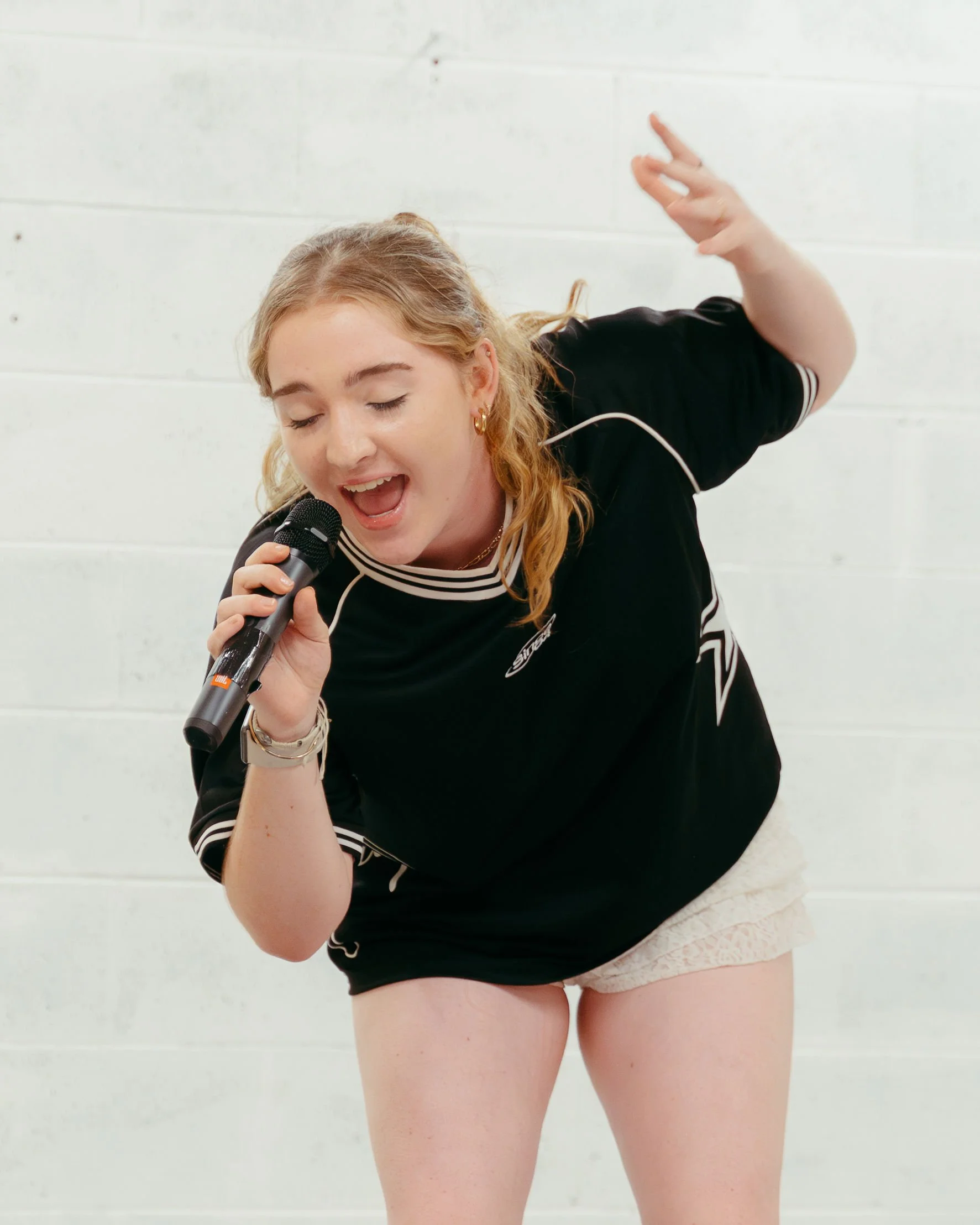 A young woman holding a microphone singing or speaking, with her eyes closed and one arm raised, against a white wall background.