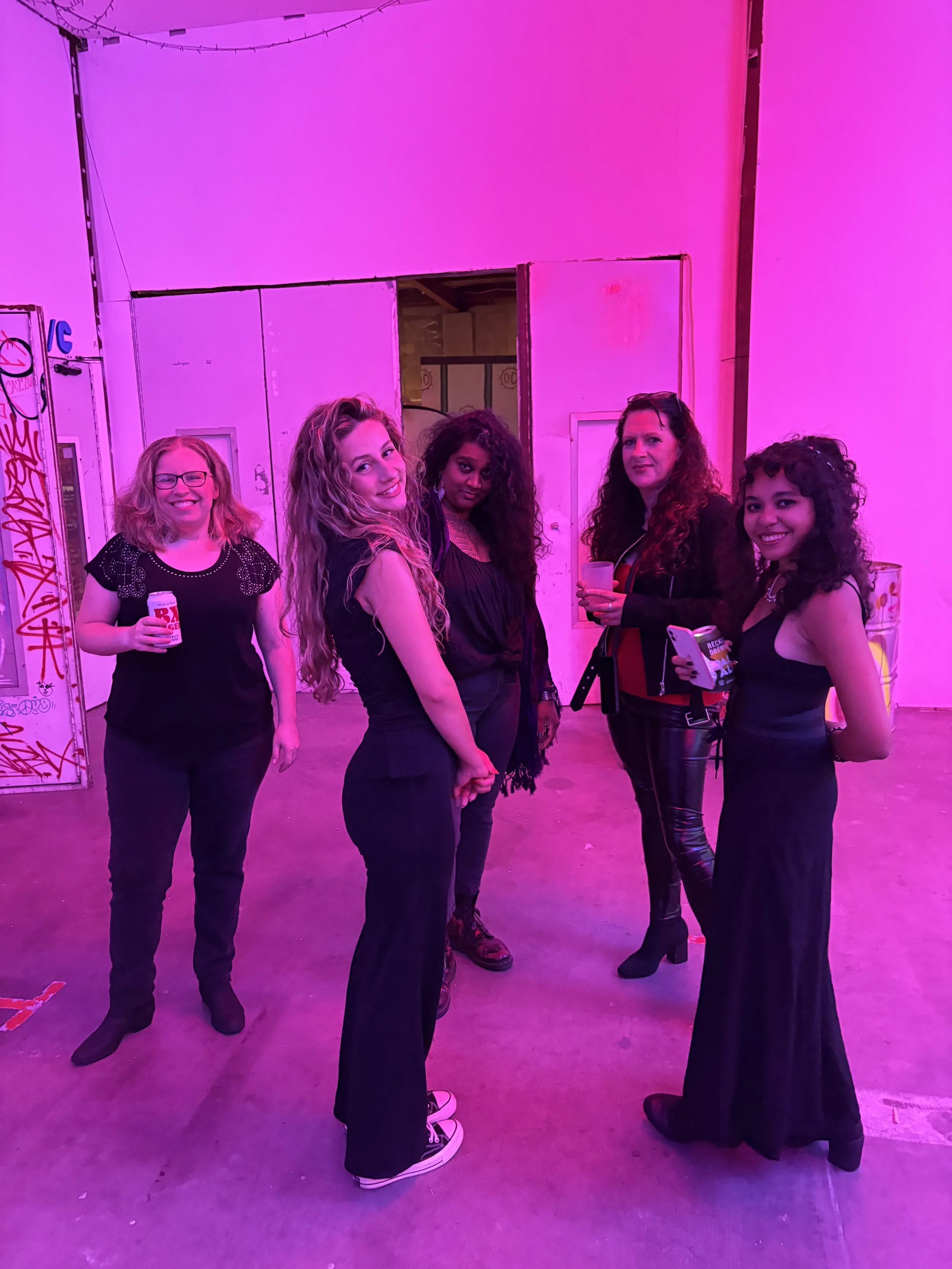 Five women standing together in a room with pink lighting, smiling and holding drinks.