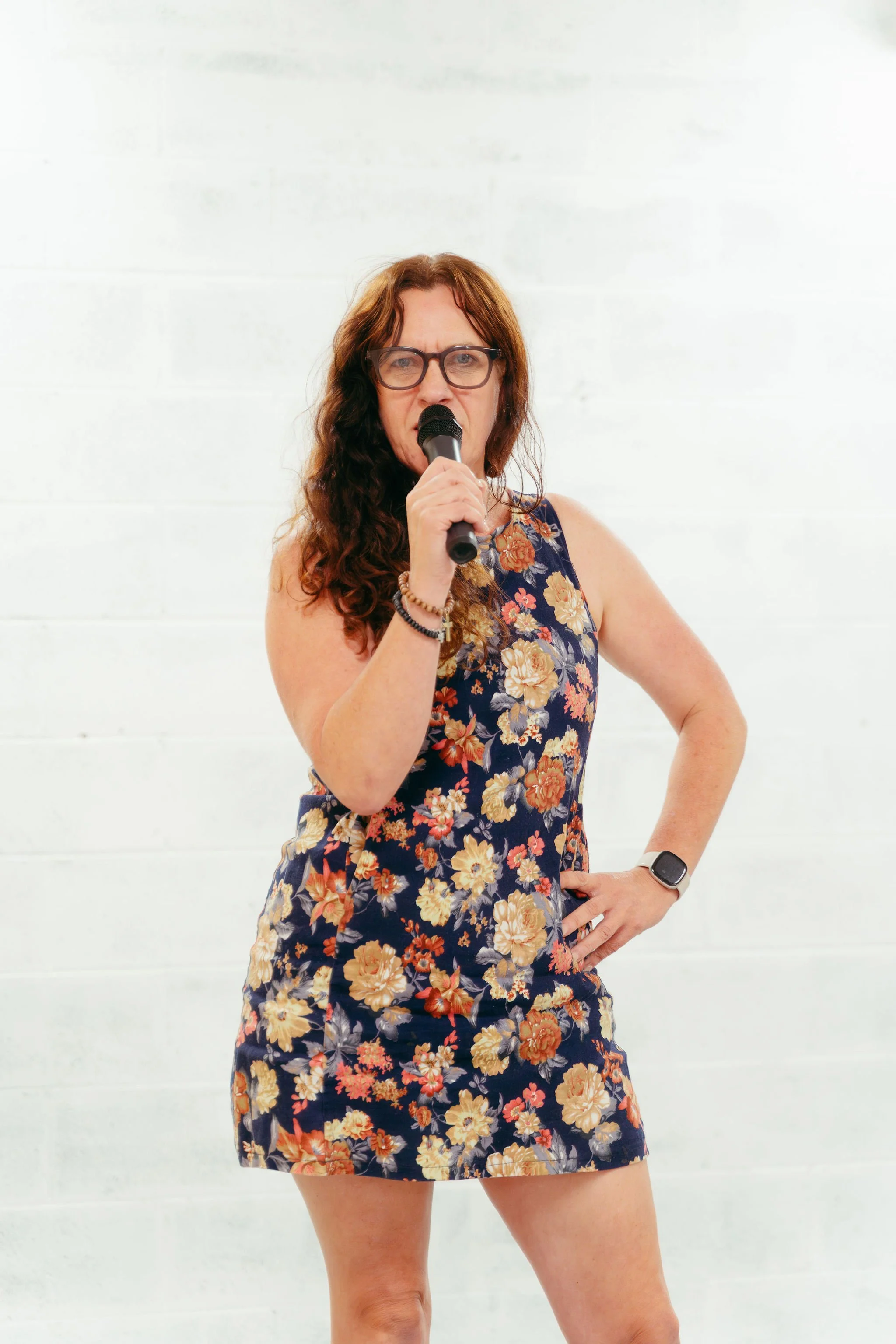 A woman with curly brown hair, glasses, and a floral sleeveless dress, holding a microphone and standing against a white wall.