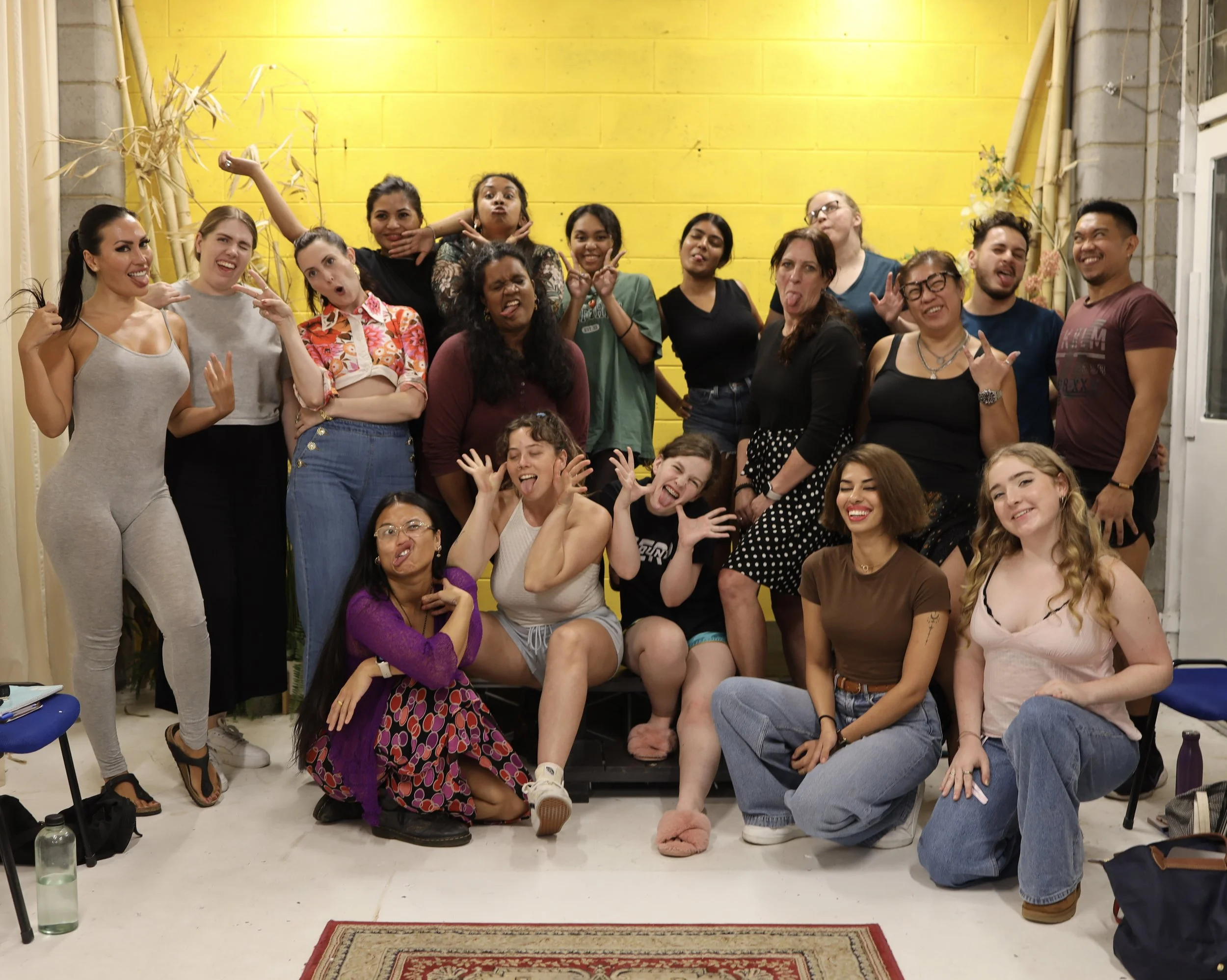 A large group of diverse people posing playfully in front of a bright yellow wall, some making funny faces and gestures.