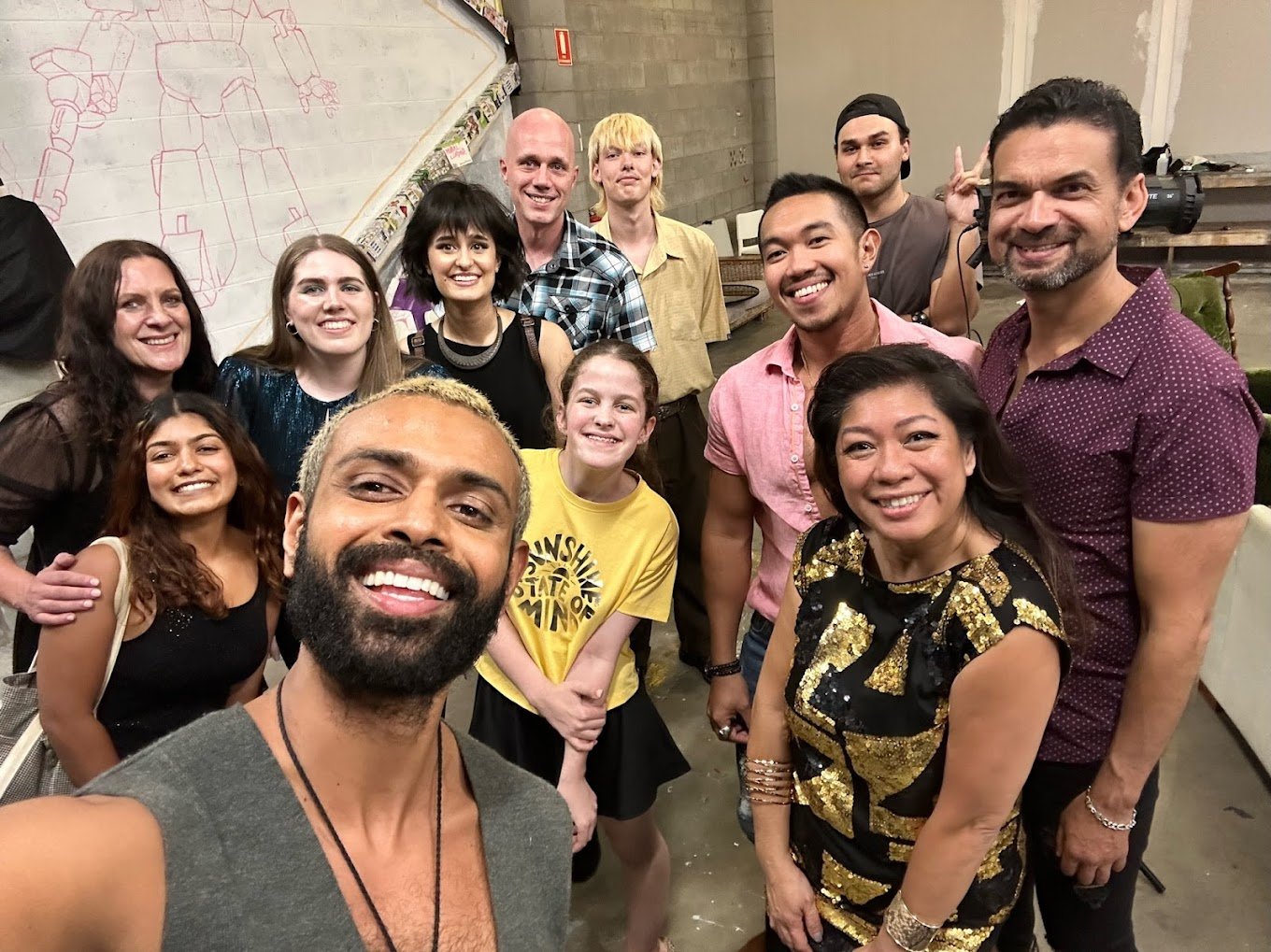 A group of people taking a selfie at an indoor event, smiling and posing in front of a wall with a sketch of a robot.