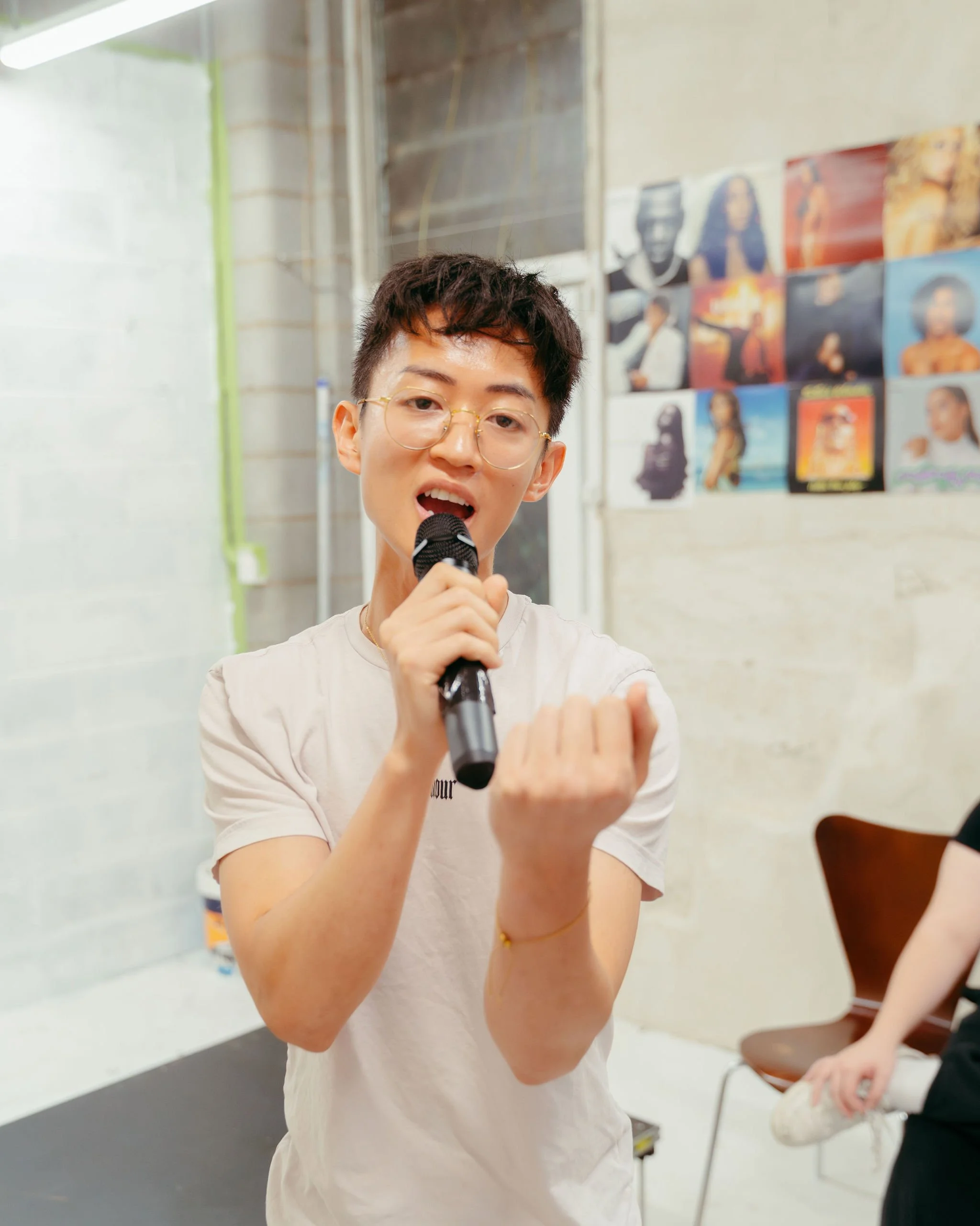 A young person with short dark hair and glasses singing into a microphone in a room with posters on the wall.