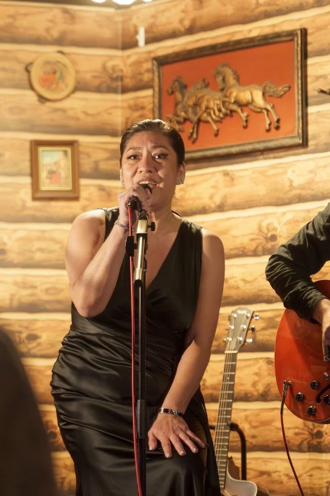 A woman singing into a microphone in a cozy, wood-paneled room with framed artwork and a guitar in the background.