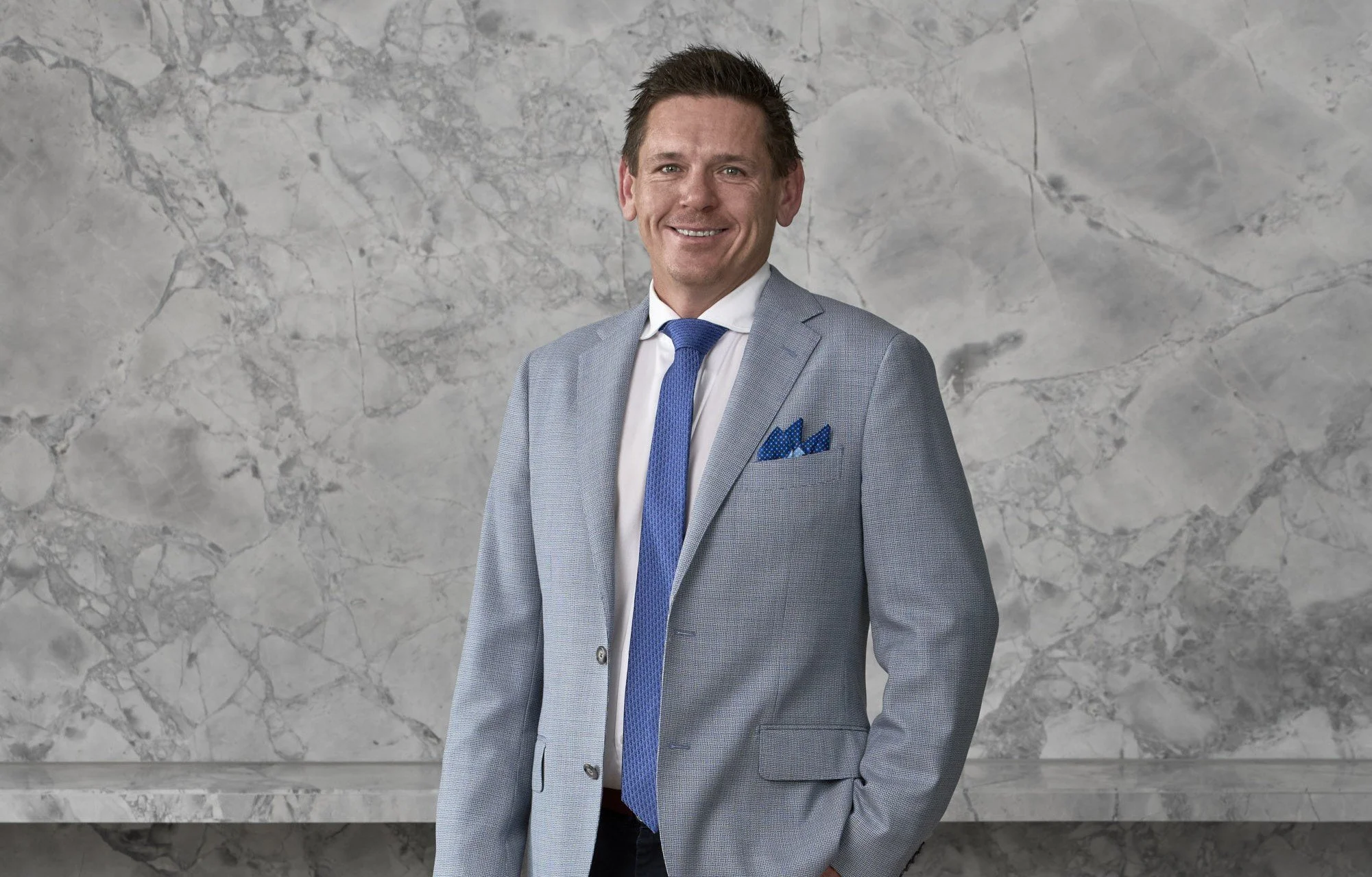 A smiling man in a light gray suit, white shirt, and blue tie, standing in front of a marble wall, with a blue pocket square in his suit jacket pocket.