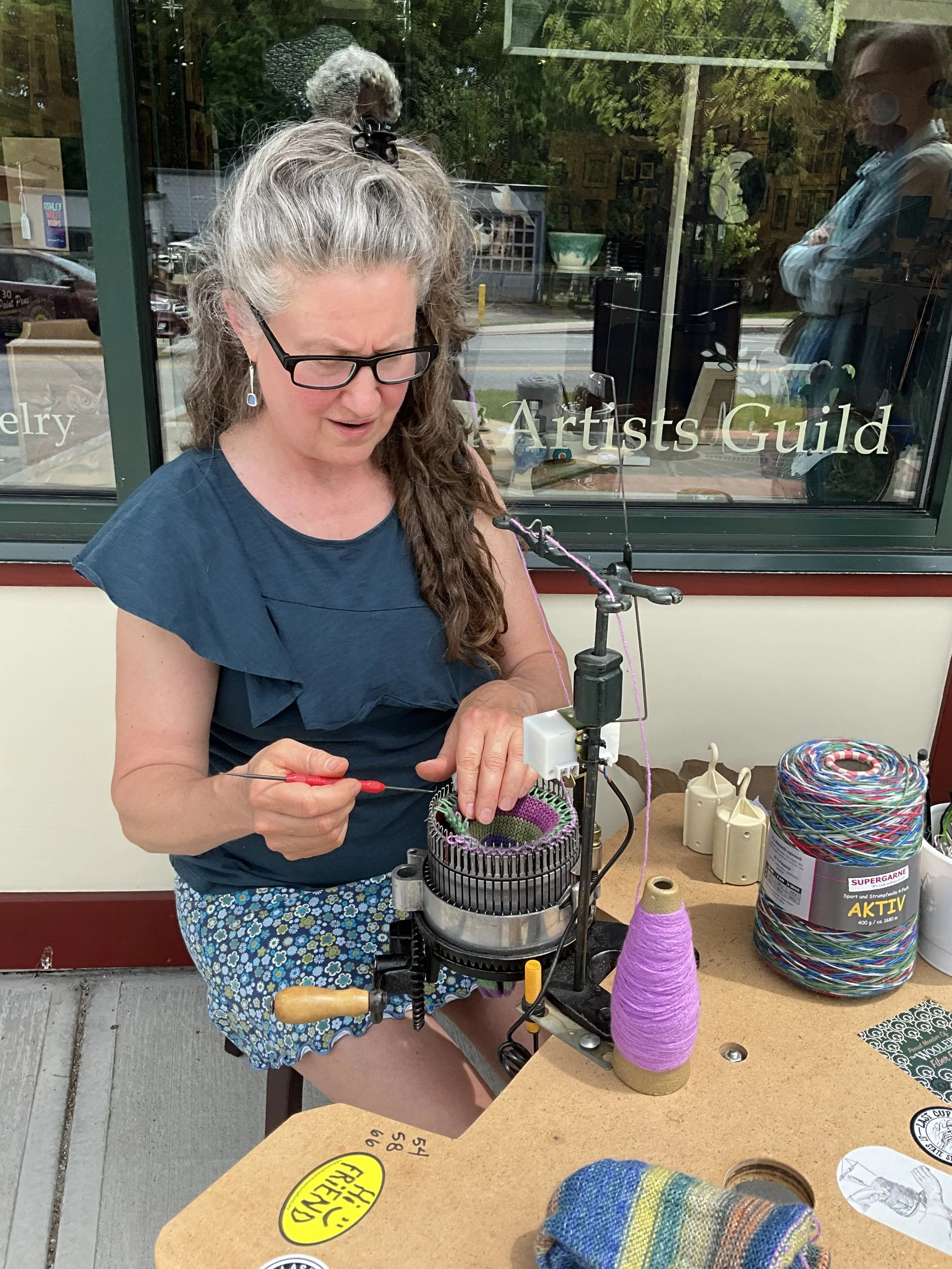 A woman with glasses and long gray hair, wearing a blue top and patterned shorts, is operating a knitting machine. She is engaged in knitting colorful yarn. There are balls of yarn, a sock, and knitting tools on the table, and the scene appears to be outside a shop window labeled 'Artists Guild.' Reflection of a person taking the photo and street view can be seen in the window.