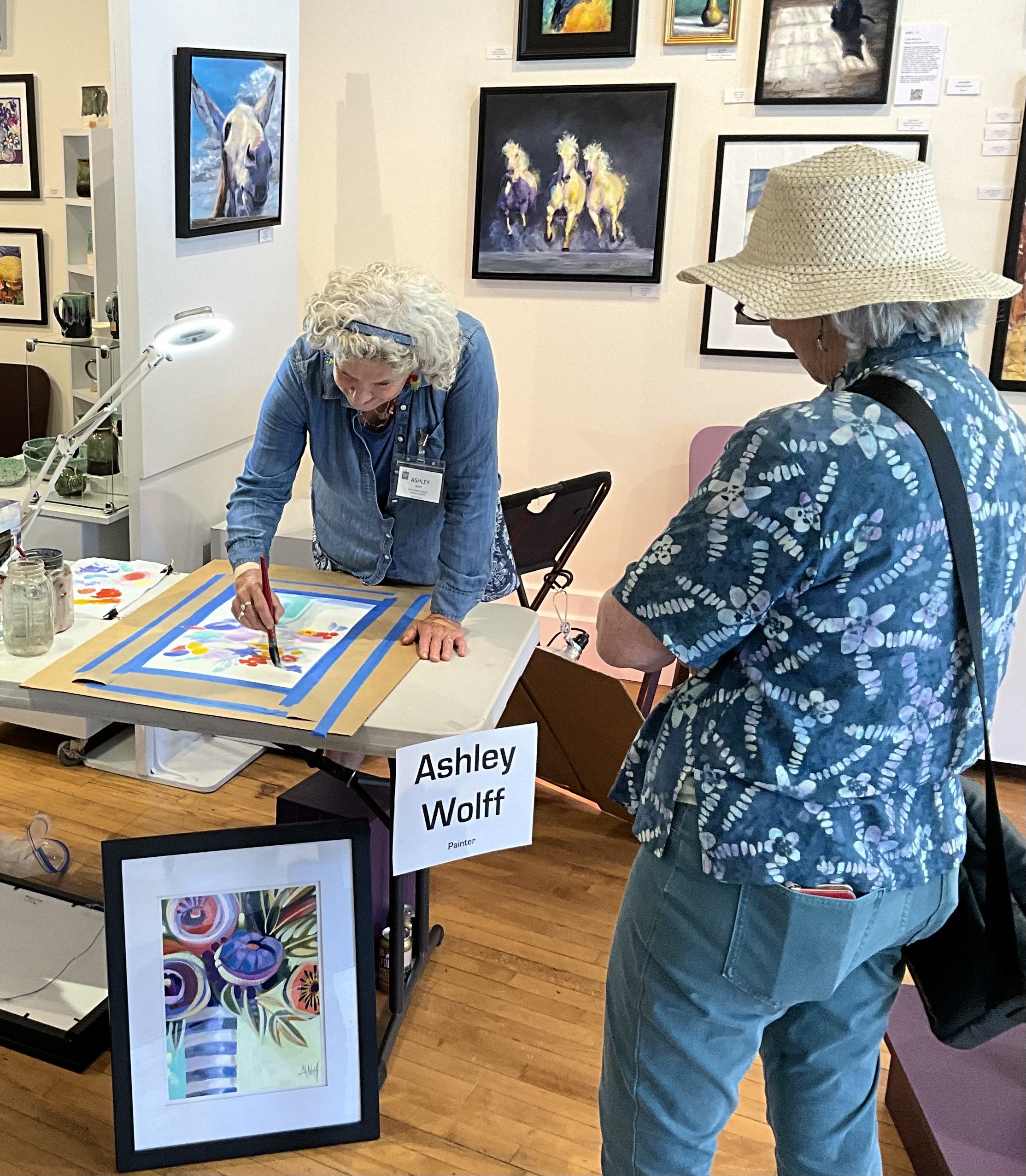 A woman with curly gray hair wearing a large wide-brimmed hat and a blue patterned shirt looks at artwork in an art gallery. Another woman with white hair and a blue shirt is painting or working on an art project at a table.