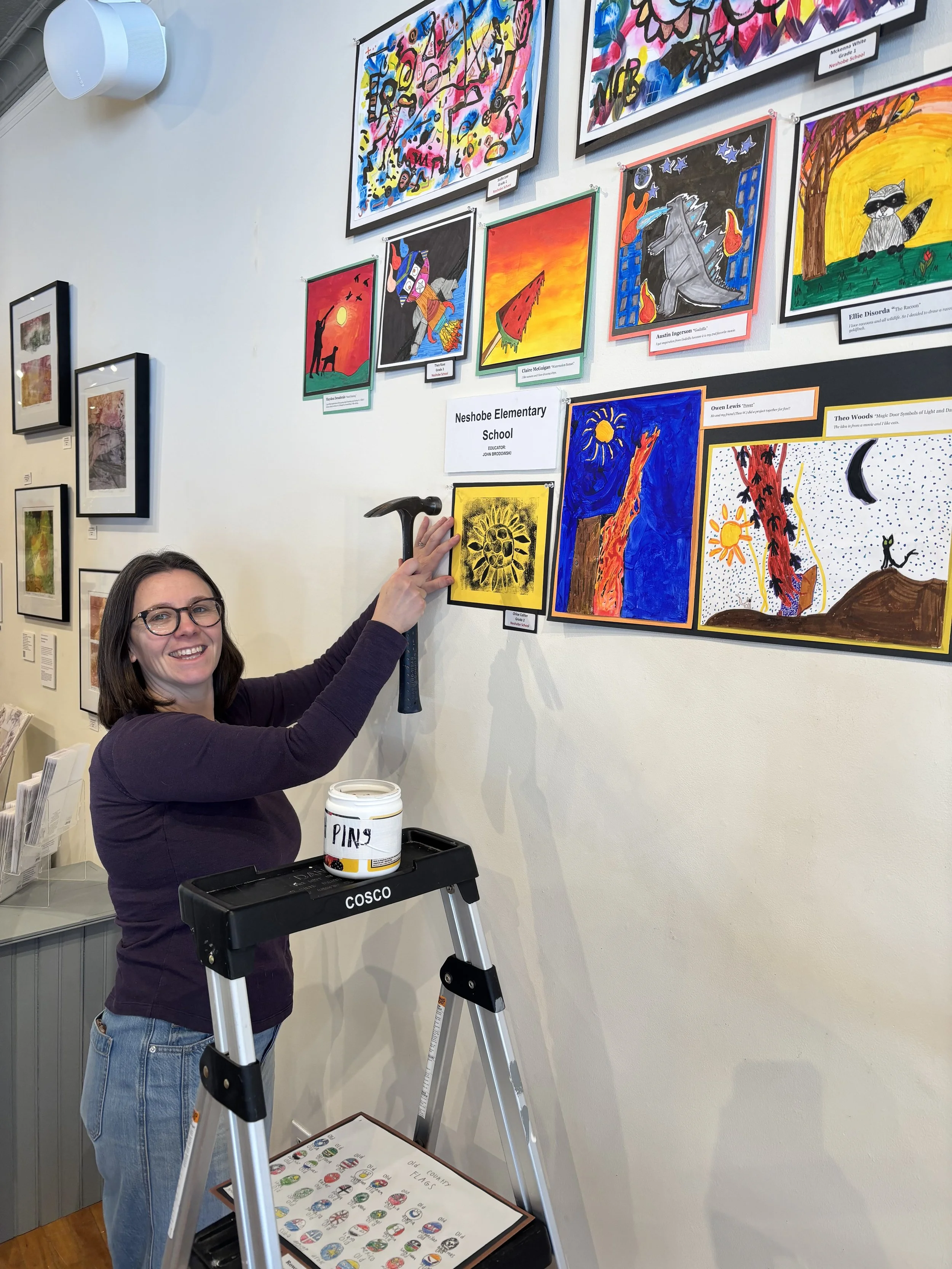 A woman wearing glasses and a purple jumpsuit is standing on a step ladder, hanging colorful artwork on a gallery wall. The artwork consists of various vibrant paintings and drawings, including a yellow sunflower with black details, a robot, a horse, and other abstract compositions. There is a white mug with the word 'PINY' on a black cart and a chart with various painted symbols on a table below.