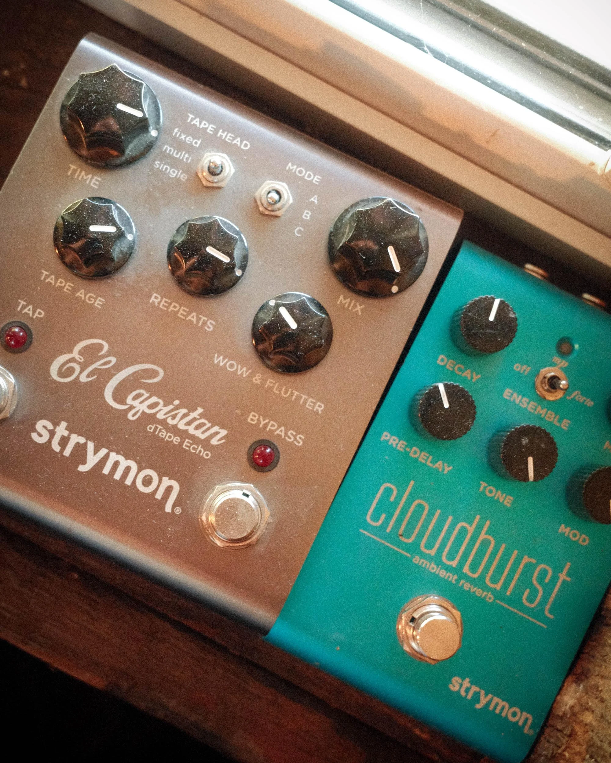 Close-up of two guitar effects pedals on a wooden surface, with knobs, switches, and labels for controls such as 'TAPE HEAD' and 'CHORUS'.