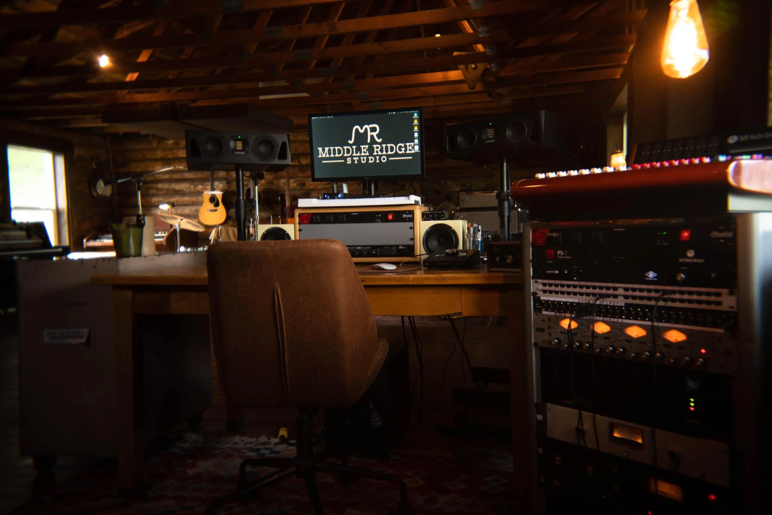A music production studio with wooden walls and ceiling, featuring a computer monitor displaying the Middle Ridge Studio logo, surrounded by speakers, a keyboard, a guitar, and audio equipment, with a window to the left letting in natural light.