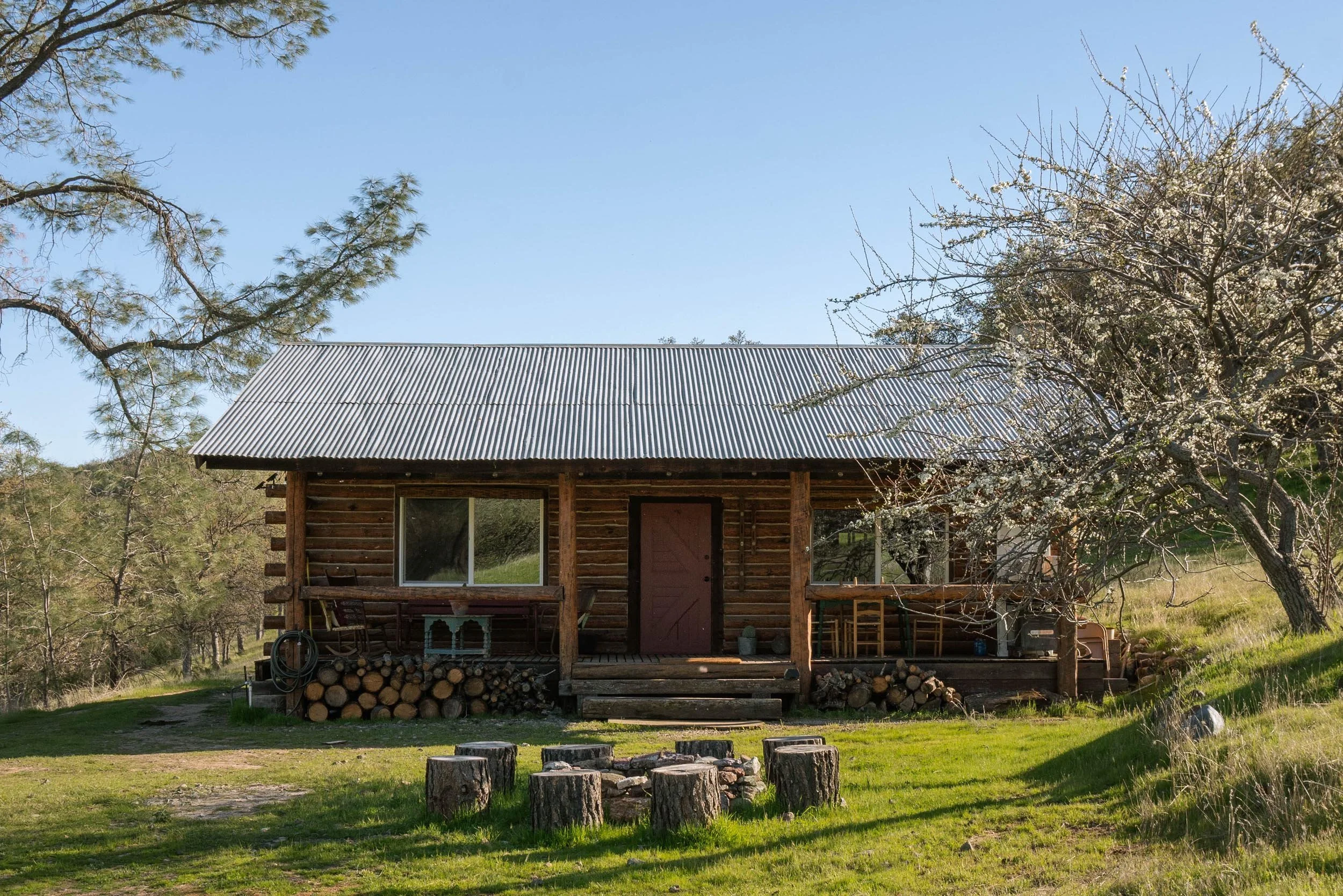 A small wooden cabin with a metal roof, set in a rural area with trees and a grassy yard. The cabin has a porch with logs stacked underneath and a window on the front. A fire pit made of logs is visible in the yard, and trees with spring blossoms sur