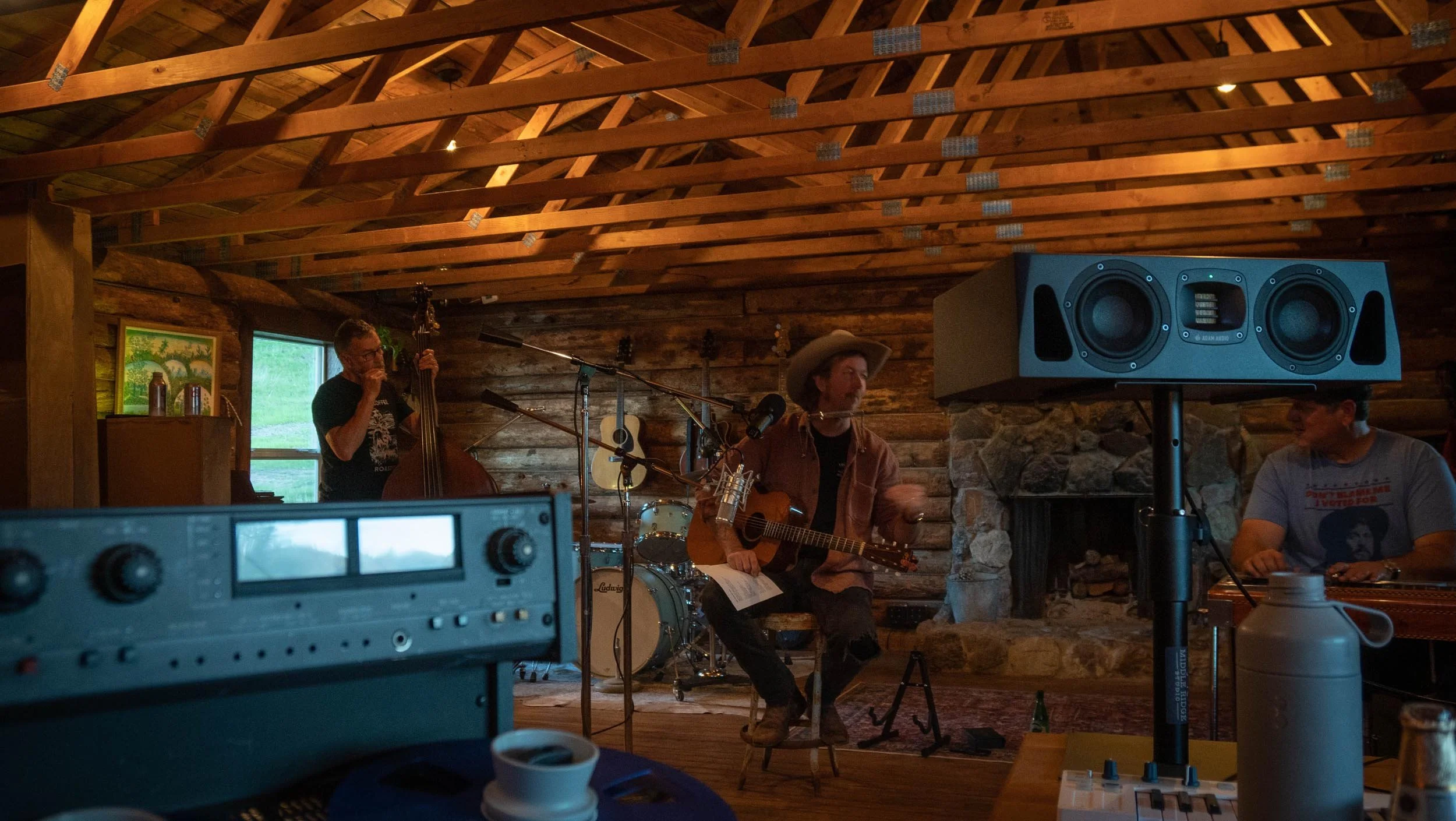 A band performing in a rustic, wooden indoor setting with three musicians: a man playing a double bass, a man singing and playing an acoustic guitar, and another man sitting at a keyboard. Large speaker, microphones, and audio equipment are visible.