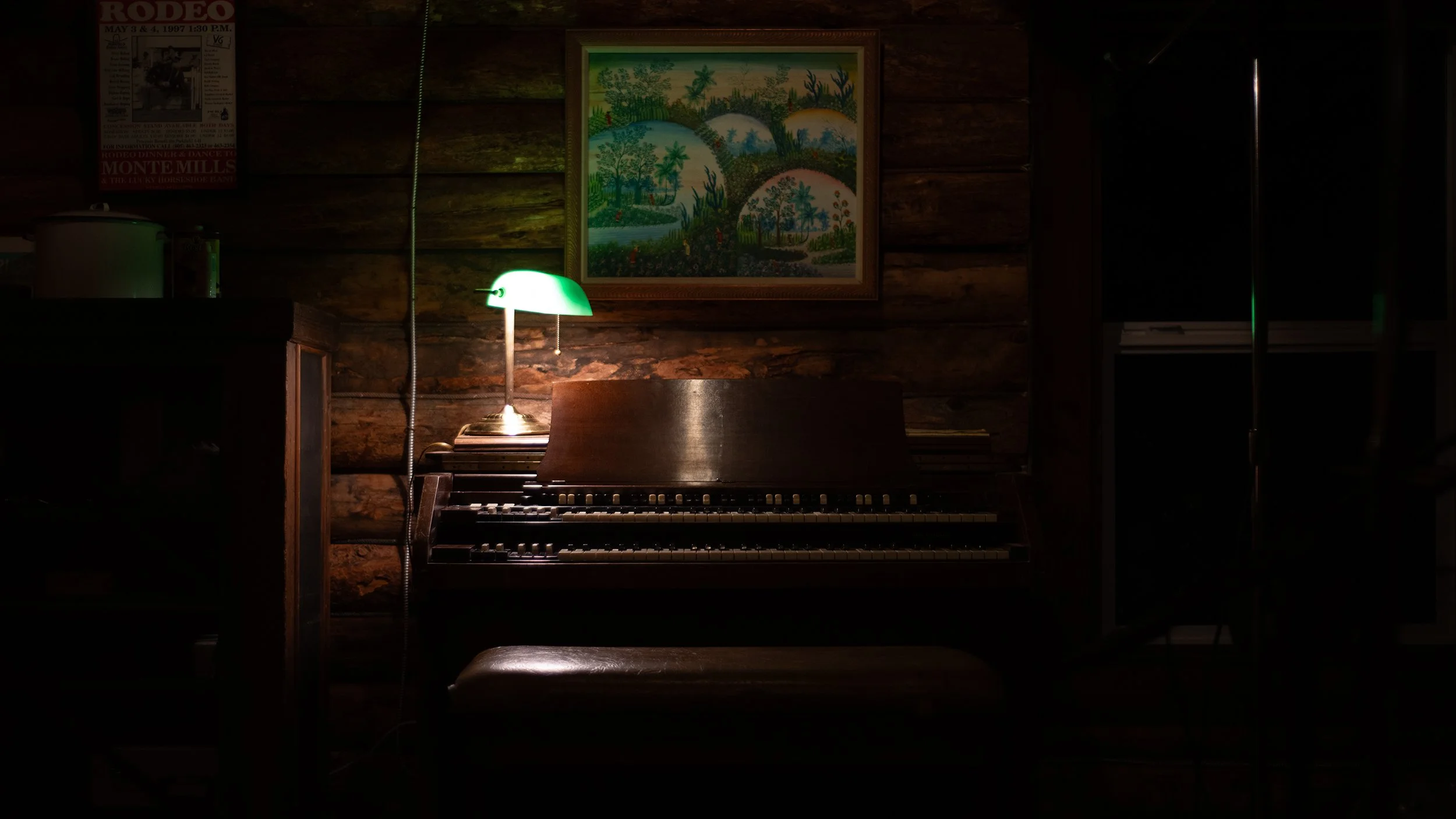 Dimly lit room with a wooden wall, a framed colorful painting, a green-shaded desk lamp, and a piano keyboard.