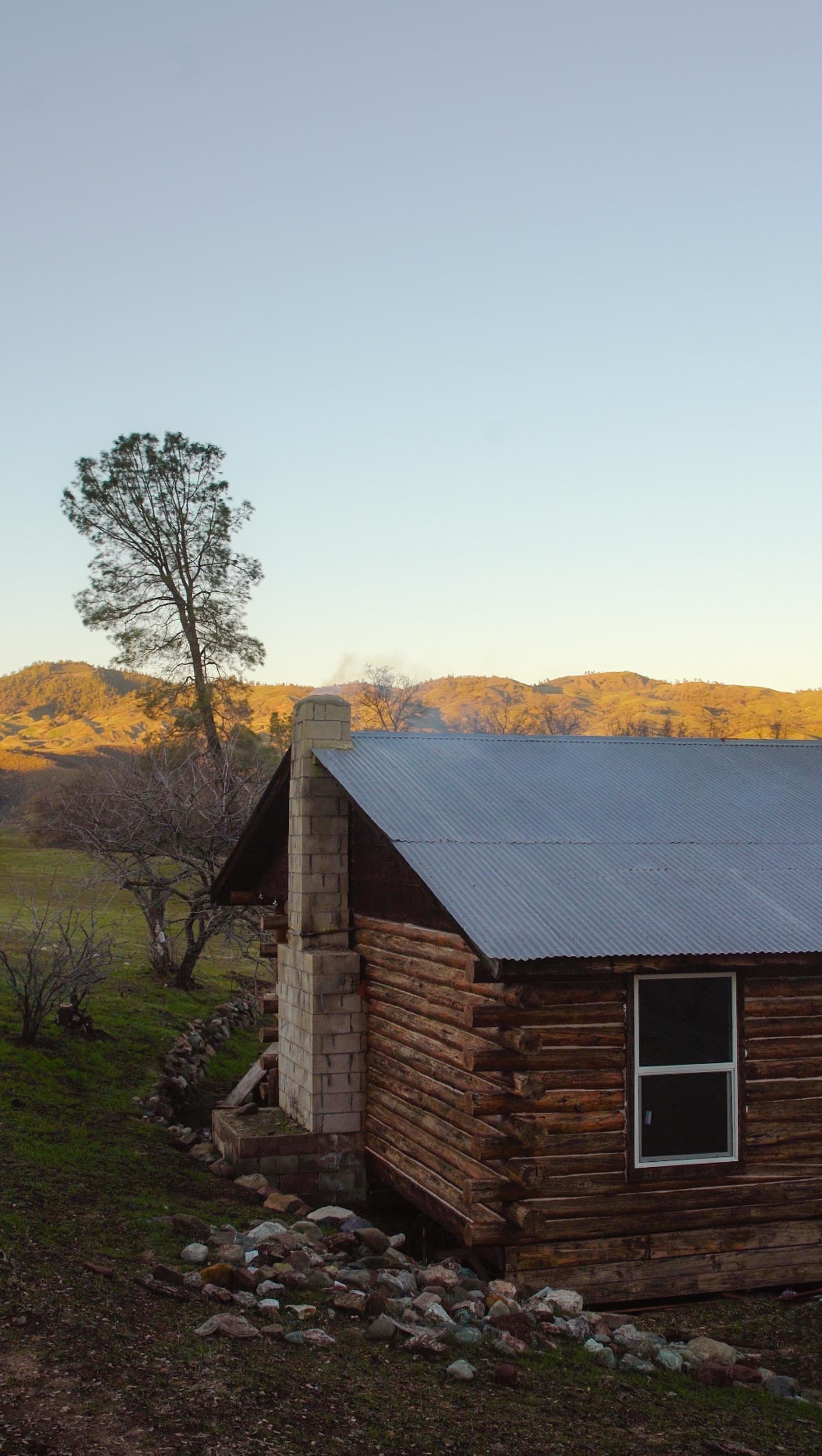 A rustic log cabin with a metal roof and chimney, surrounded by trees and rocks, set against rolling hills and a clear sky at sunset.