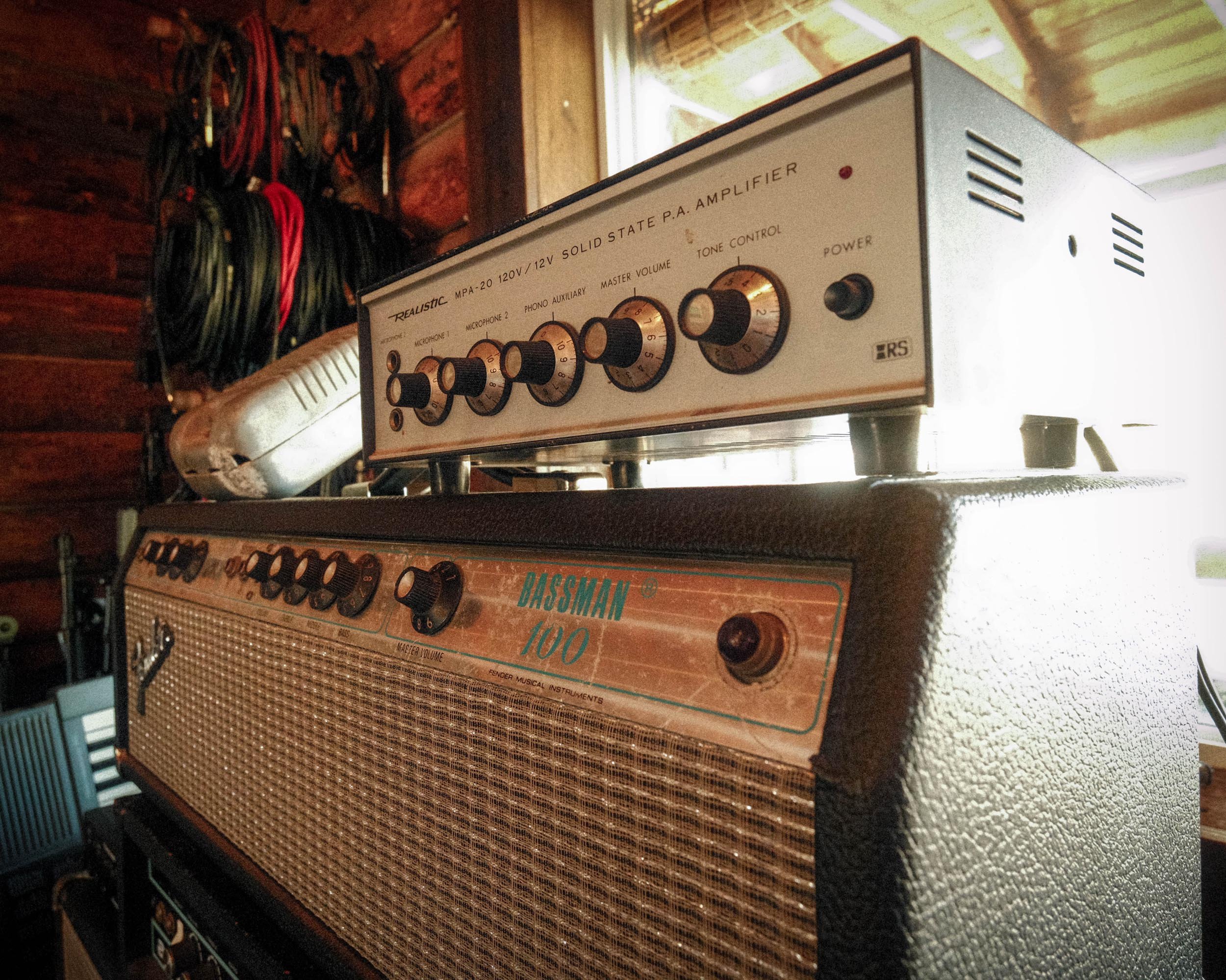 An image showing a vintage solid state P.A. amplifier with several control knobs on the front panel, placed on top of a larger speaker cabinet. Behind the amplifier, there is a wall with multiple audio and power cords hanging on hooks. Natural light 