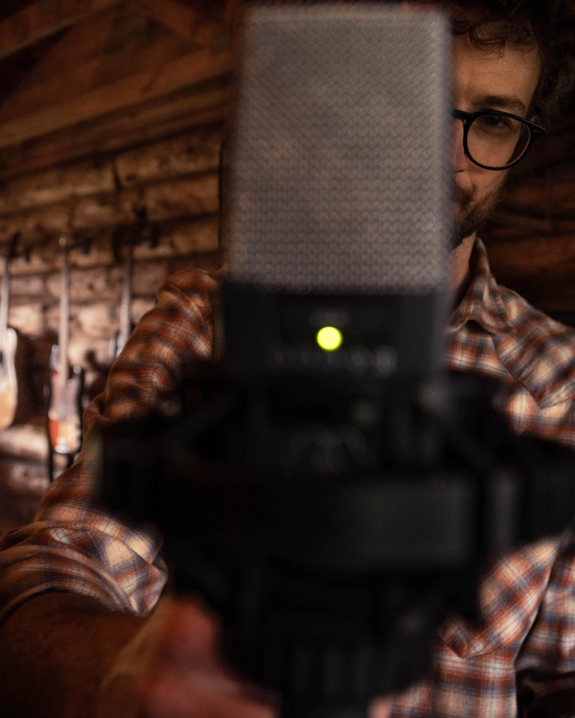 A man with curly hair, glasses, and a beard taking a selfie with a smartphone, partially obscuring his face. He is wearing a plaid shirt and standing in front of a background with wooden racks.