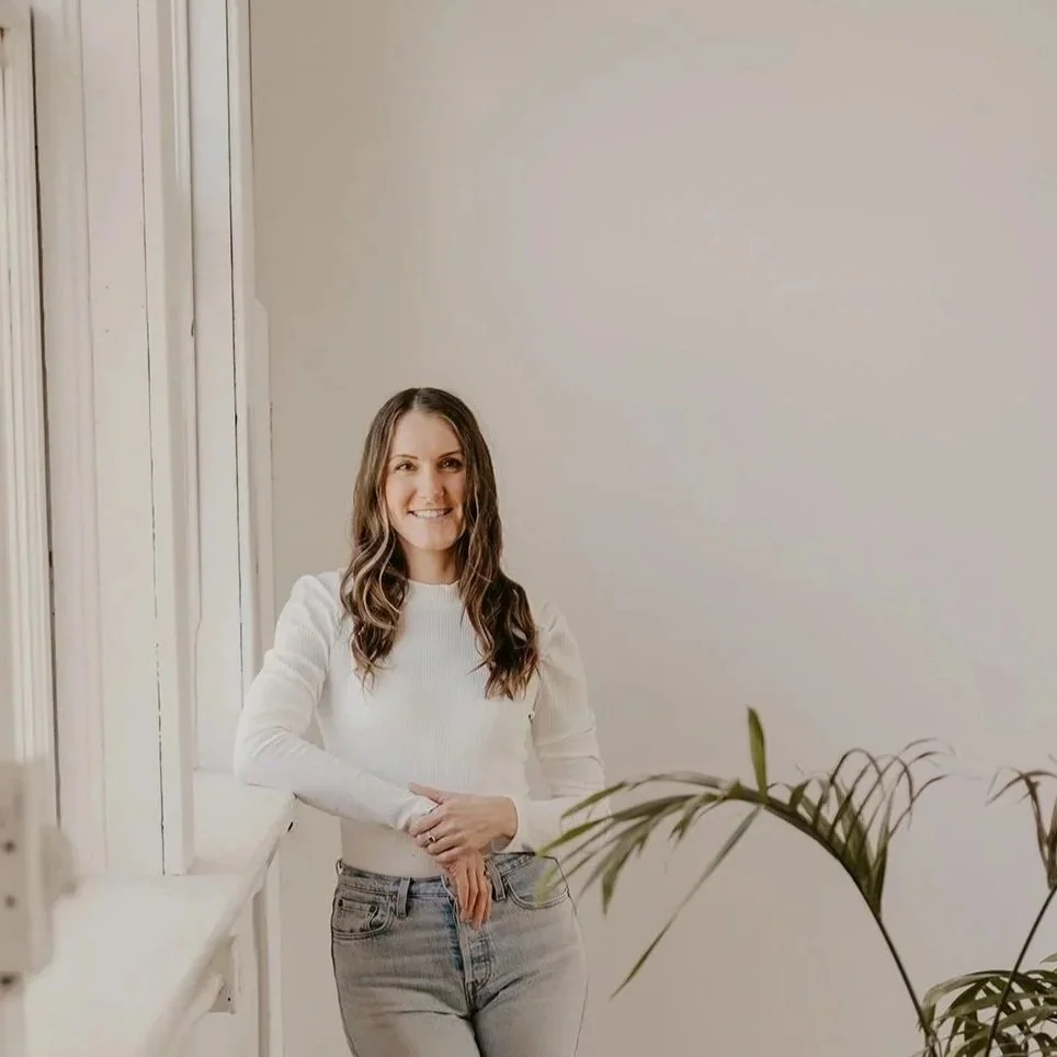A homeopath  standing next to a window with natural light and a potted plant partially visible in the foreground.