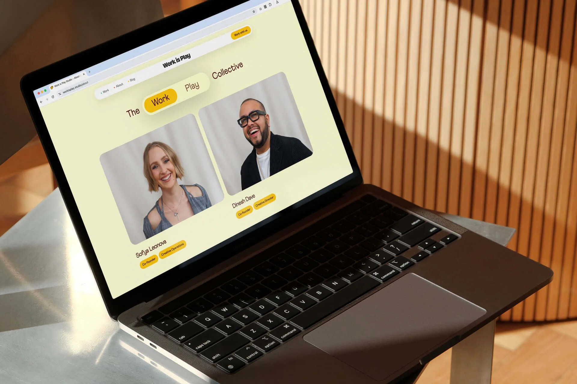  A laptop displays a website with a yellow background. The page shows two photos: one of a woman labeled "Sofya Leonova" and one of a man labeled "Dinesh Dave." The website tabs read "The Work Play Collective" with the ability to toggle between "Work