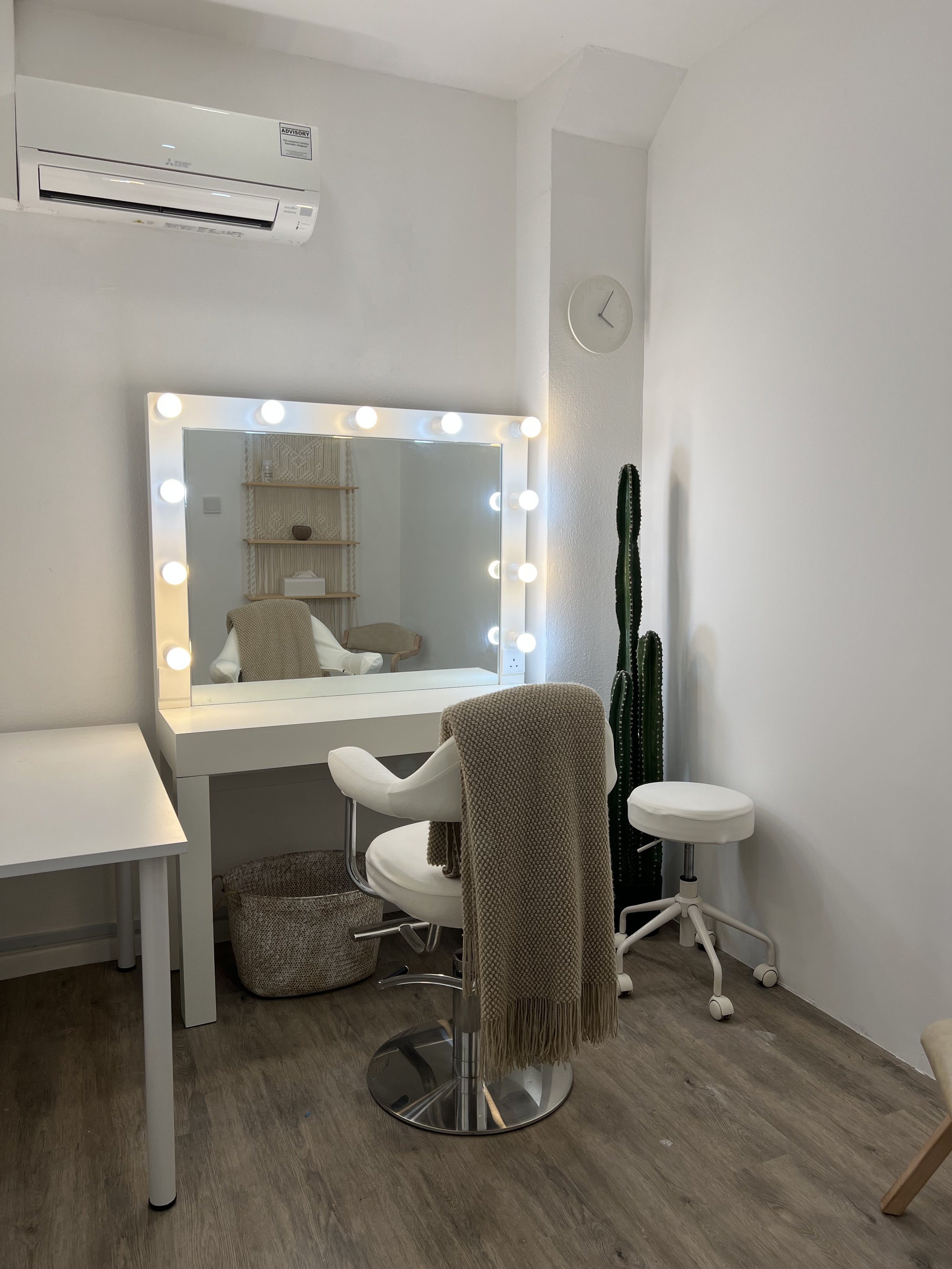 A makeup or dressing room with a illuminated mirror, a white chair with a brown throw, a tall cactus, and a white stool, all against white walls with a wood floor.