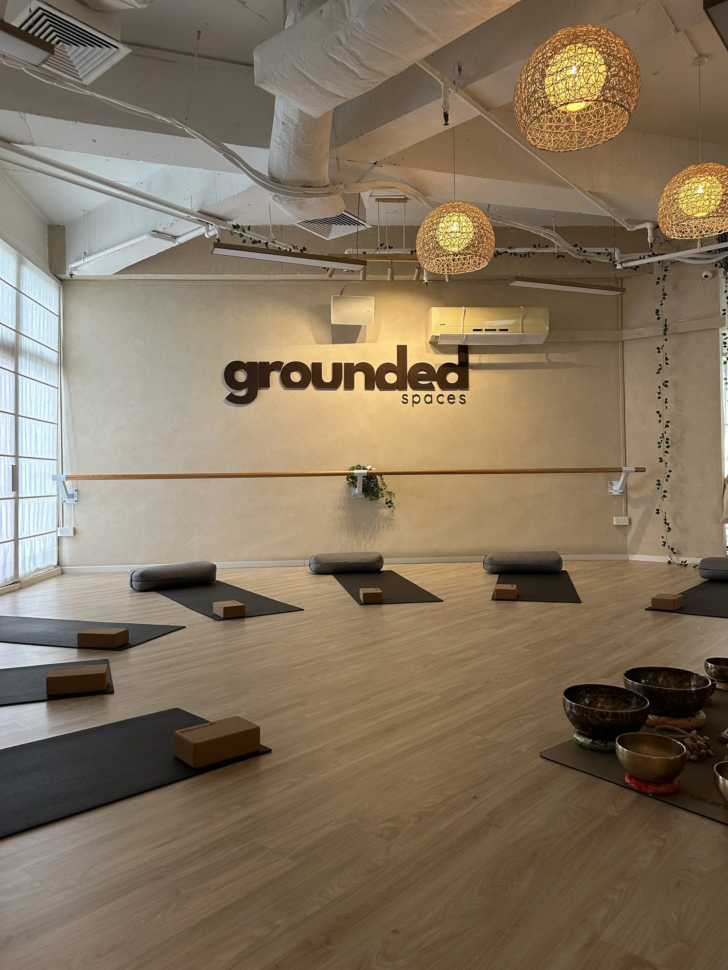 A yoga studio with black mats, bolsters, and blocks arranged in a line on a light wooden floor. The studio has a beige wall with the words 'grounded spaces' displayed. Hanging woven light fixtures and a ballet barre along the wall are visible, with some greenery and natural light from windows.