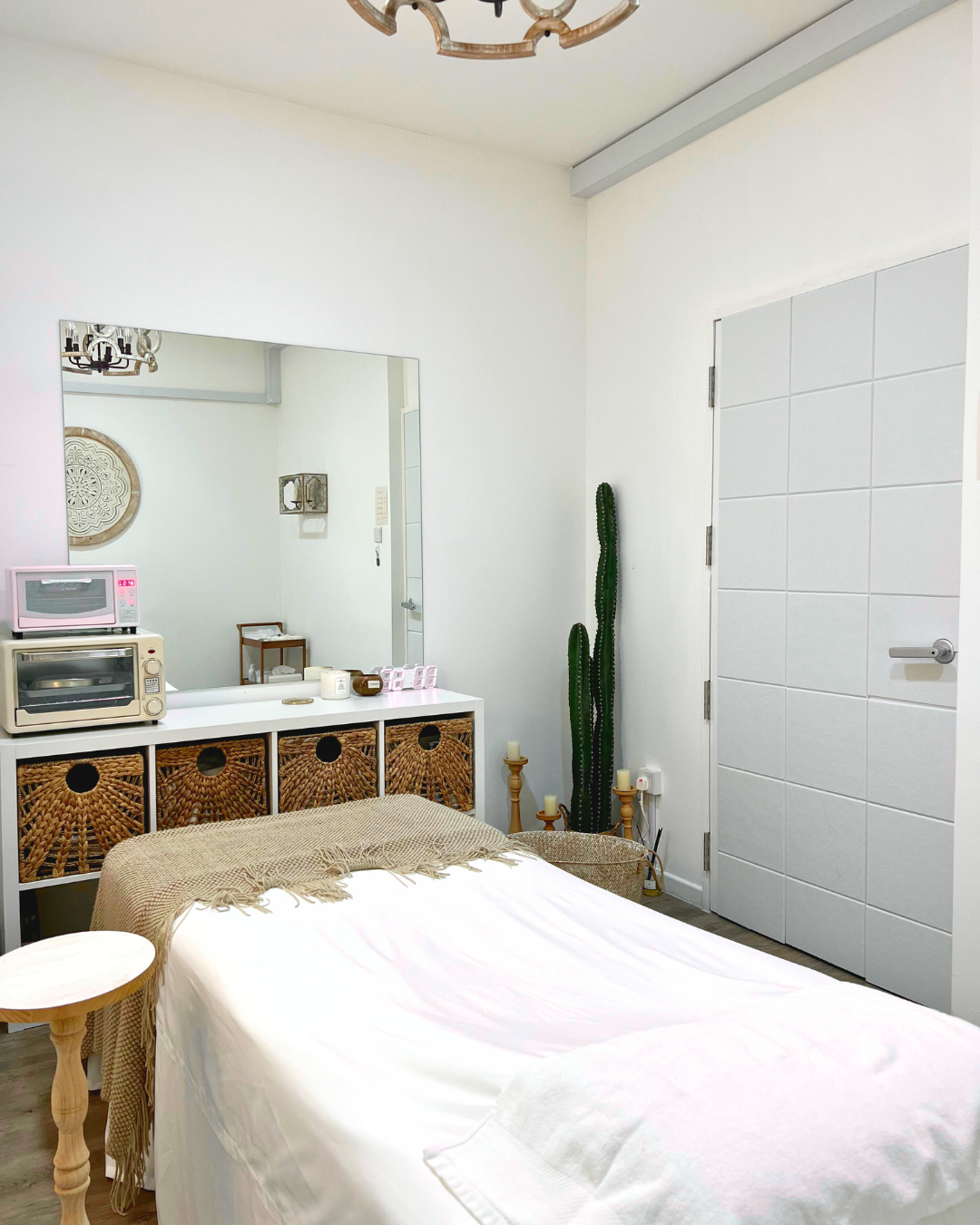 A bedroom with a bed covered in white sheets, a woven wicker headboard, and a beige throw blanket, a white side table, a large mirror, a cactus plant, a white door, and various decorative items on the dresser, including a microwave and a toaster.