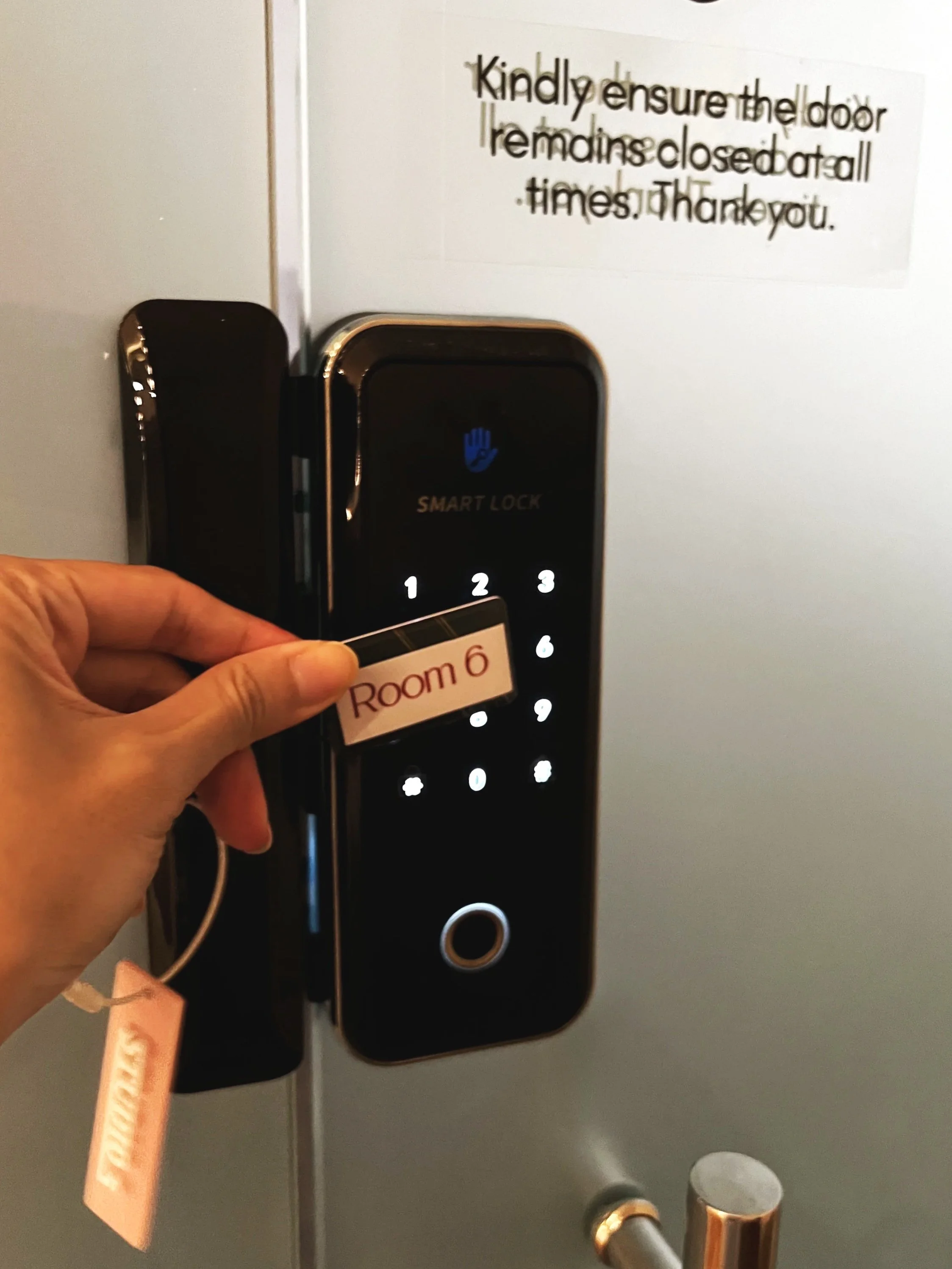 Close-up of a black smart lock door lock with a person holding a Room 6 keycard in front of it. There is a sign on the door with instructions to keep the door closed at all times, thanking the guest.
