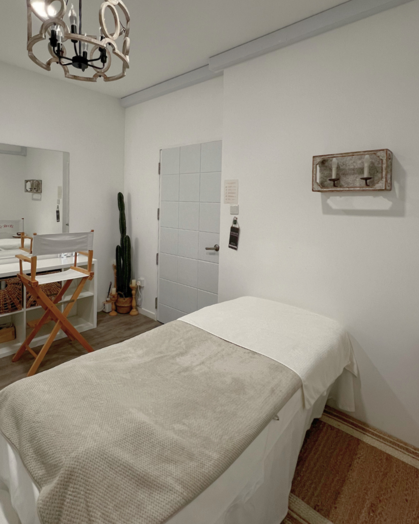 A cozy, modern bedroom with white walls and wooden flooring, featuring a bed with white linens, a black ring light on a stand, a mirror on the wall, a tall cactus plant, a white storage unit with woven baskets, and decorative items including a chandelier and a wall shelf.