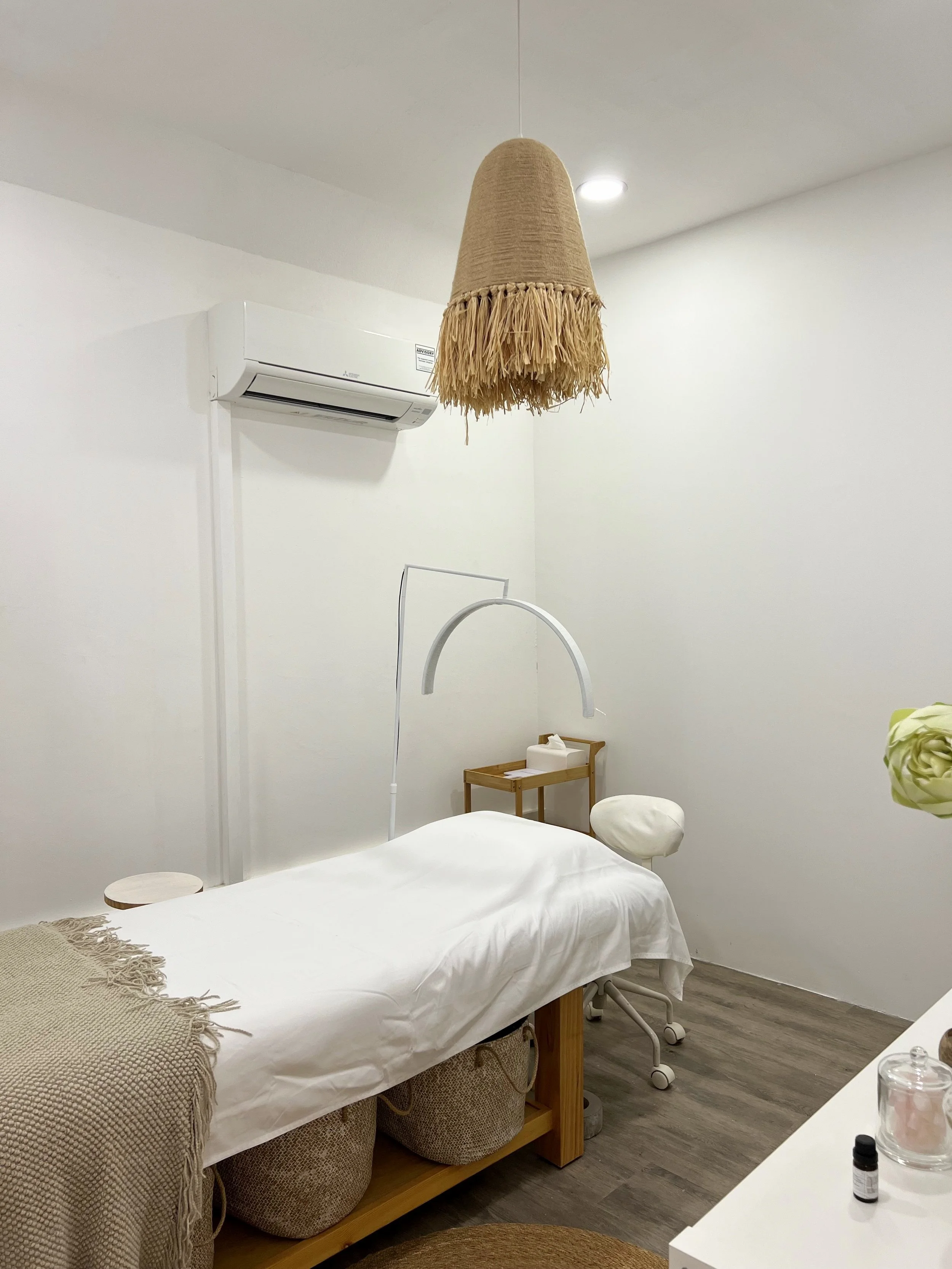 A minimalist massage or therapy room with a white treatment table, a woven beige cover, and baskets underneath. There is a hanging fringed lampshade, an air conditioner on the wall, a small side table with tissues, and a white adjustable lamp arching over the table. The room has white walls and wood-like flooring.