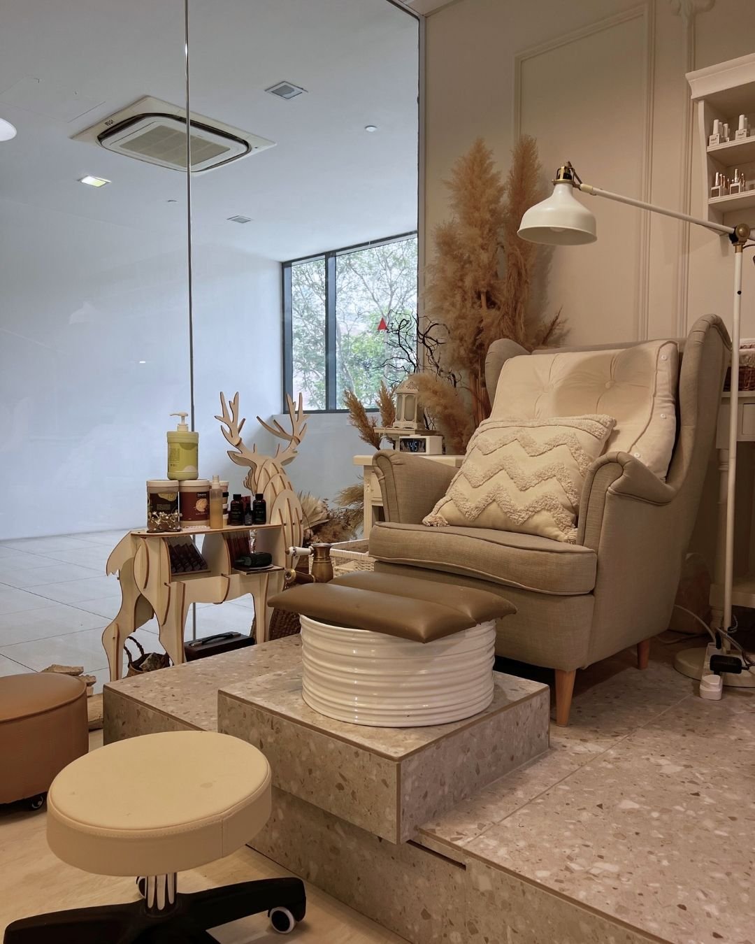 A cozy indoor space with neutral-colored furniture, including an armchair with a cushion, a small rolling stool, and a decorative large ceramic pot. There is a wooden reindeer sculpture and dried decorative plants behind the armchair, with a desk lamp attached to the wall, and a window with a view of tree outside. The overall decor is warm and minimalistic.