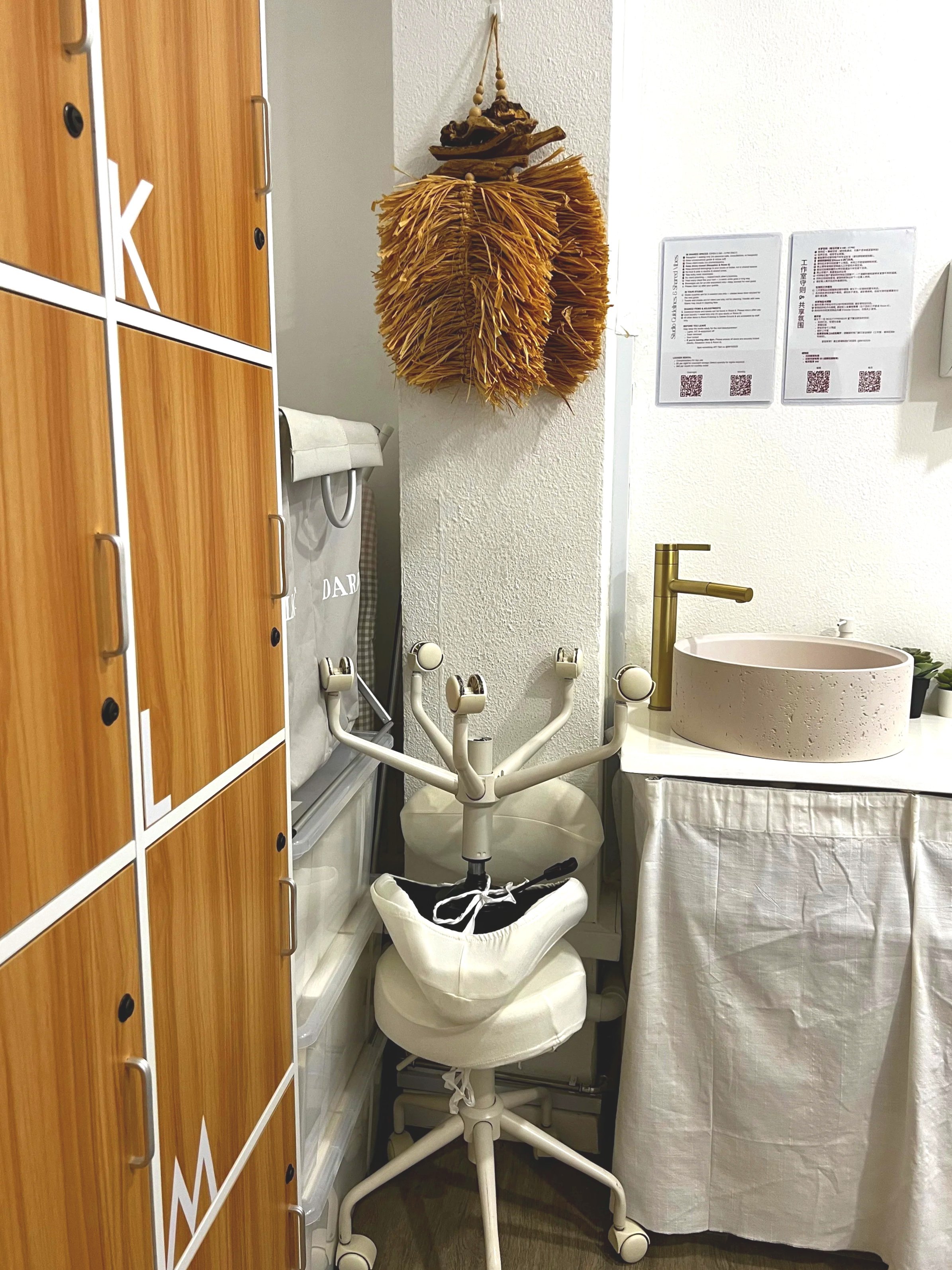 A small corner of a room with a wooden cabinet on the left, an office chair with a cloth covering the seat and a white headrest, a hanging dried plant or decorative item on the wall, a white sink with a gold faucet on a table with a white cloth, and some posters on the wall.