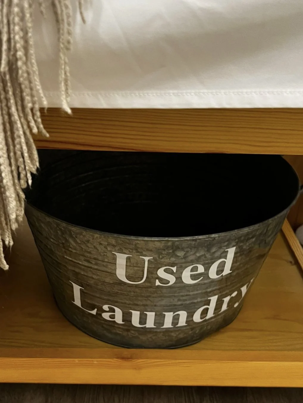 A black laundry basket with the words "Used Laundry" printed on it, placed under a wooden shelf with a white fabric above.
