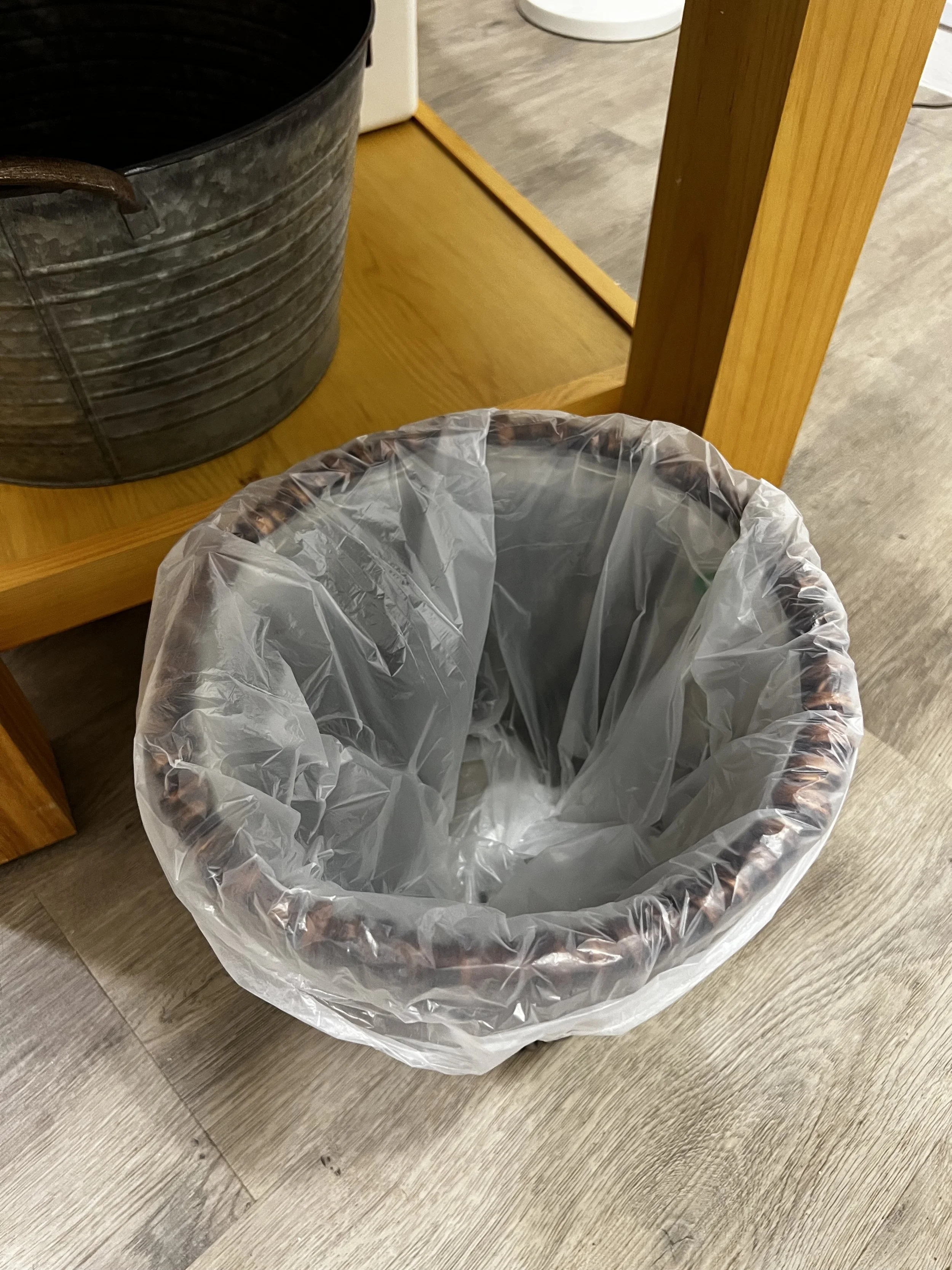 A plastic trash can liner inside a round, brown wicker trash can, placed on a wooden floor.