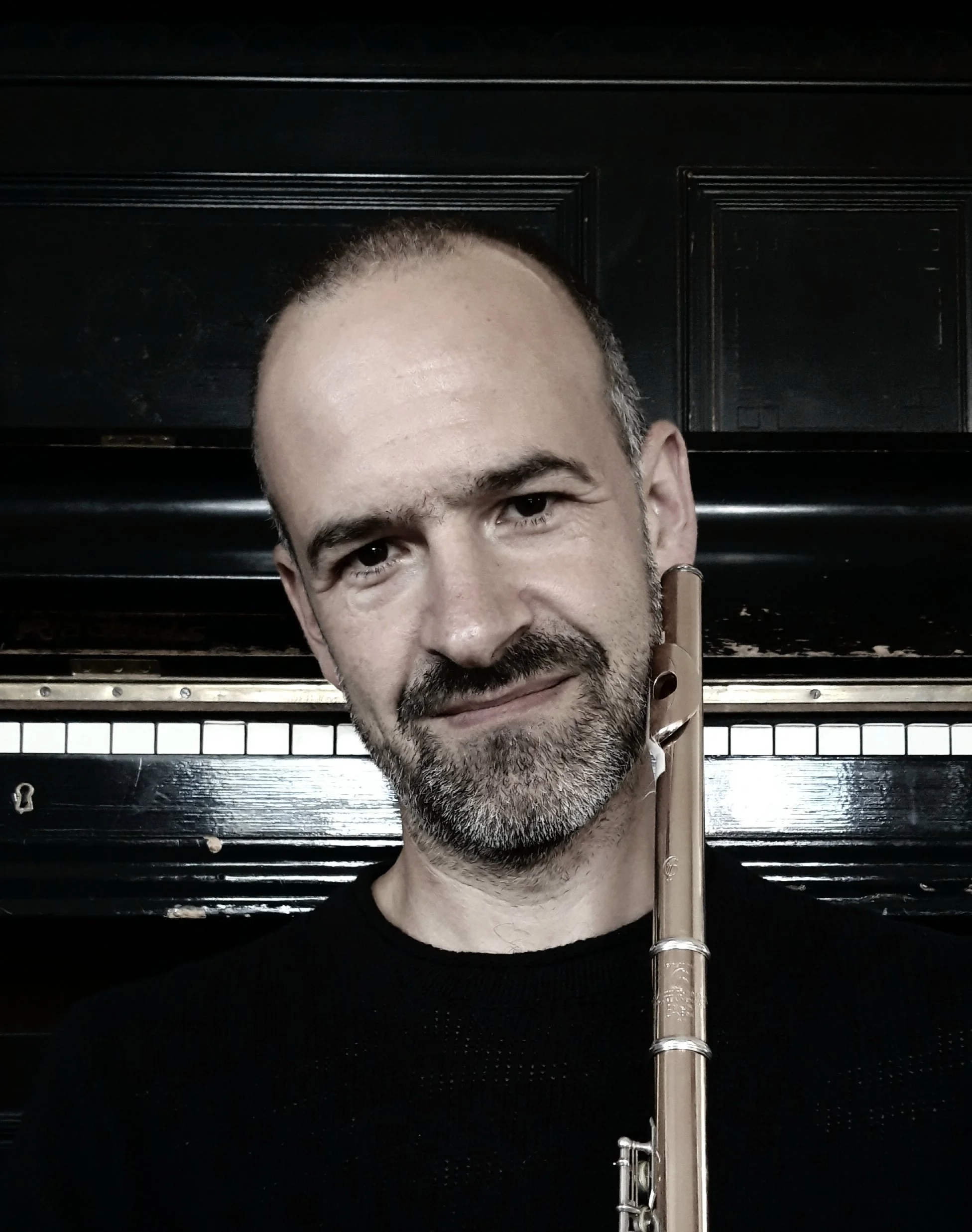 A man smiling with a wooden flute resting on his shoulder, wearing a navy blue T-shirt.
