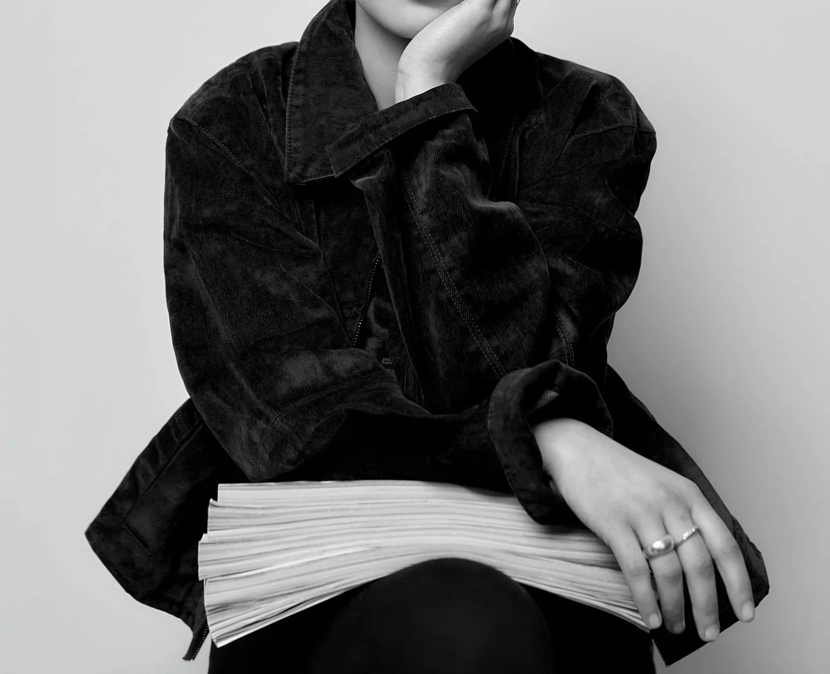 A person wearing a dark jacket, sitting with arms crossed, resting on a closed book or stack of papers. The person is not fully visible, with only part of their face, hand, and lap visible in black and white.