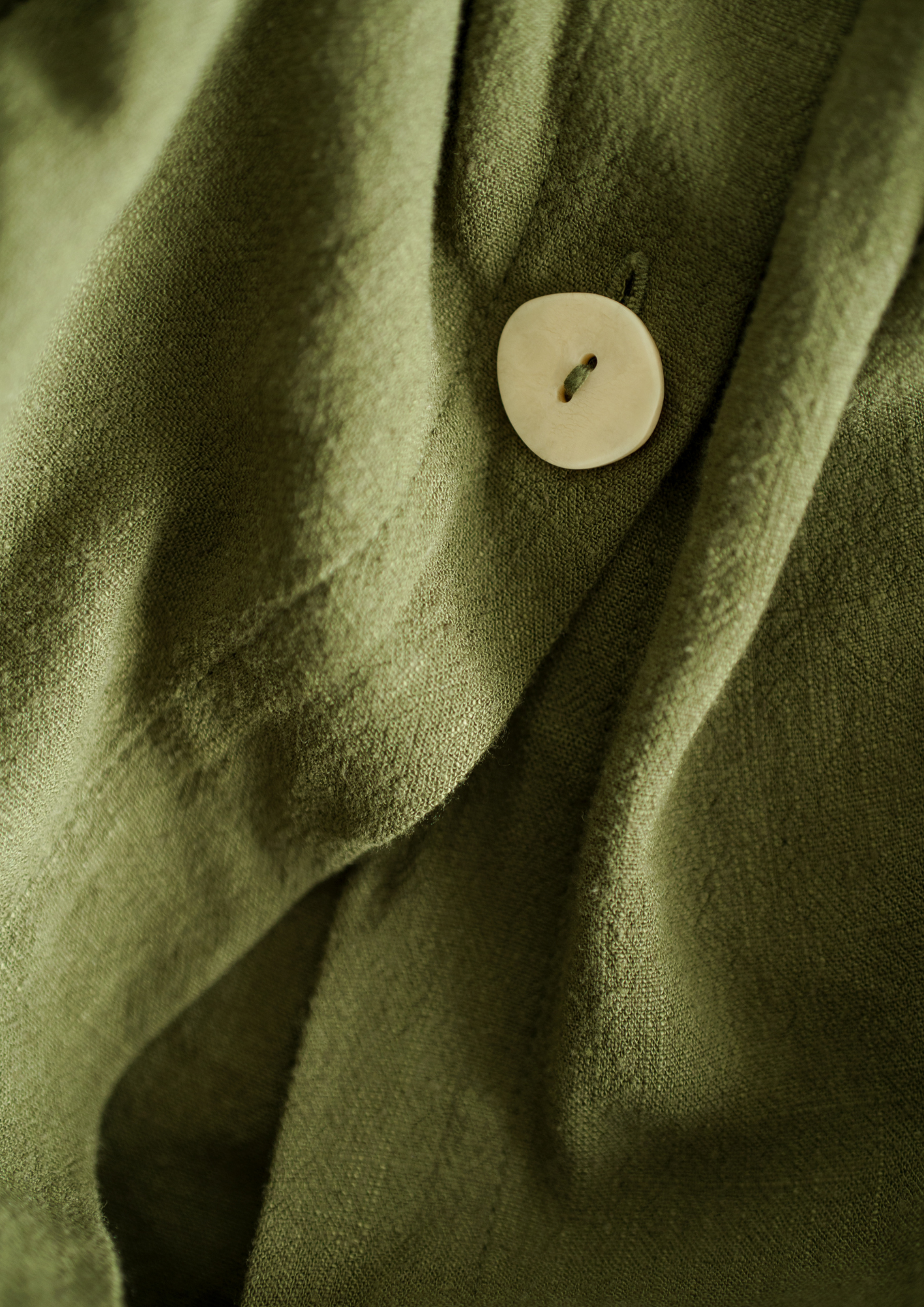 Close-up of an olive green linen shirt with a cream button, showing the weave of the fabric. Design Mood by ARDISENOSTUDIO