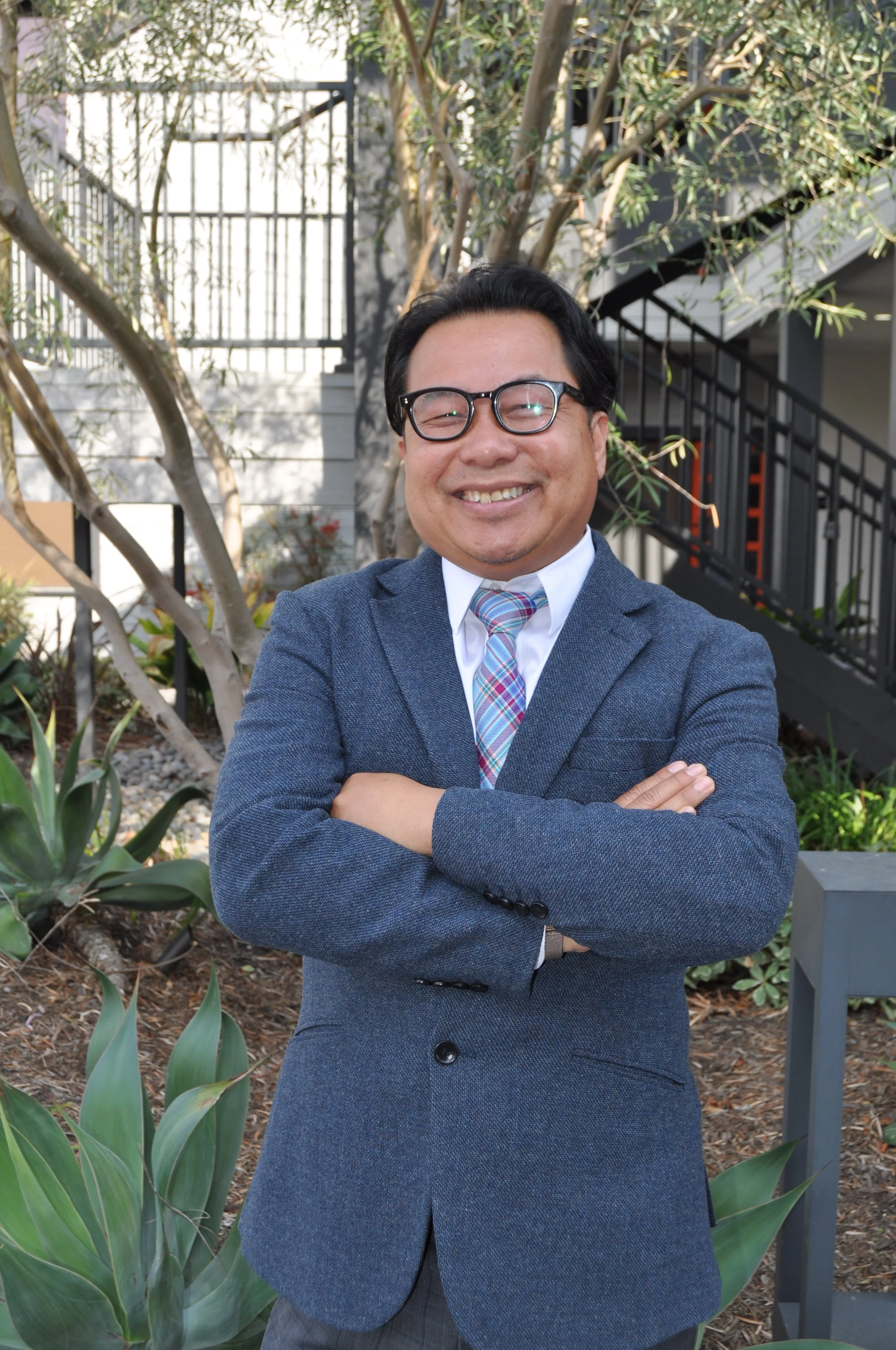 A man in a gray suit, white shirt, and colorful plaid tie smiling outdoors with trees and a building in the background.