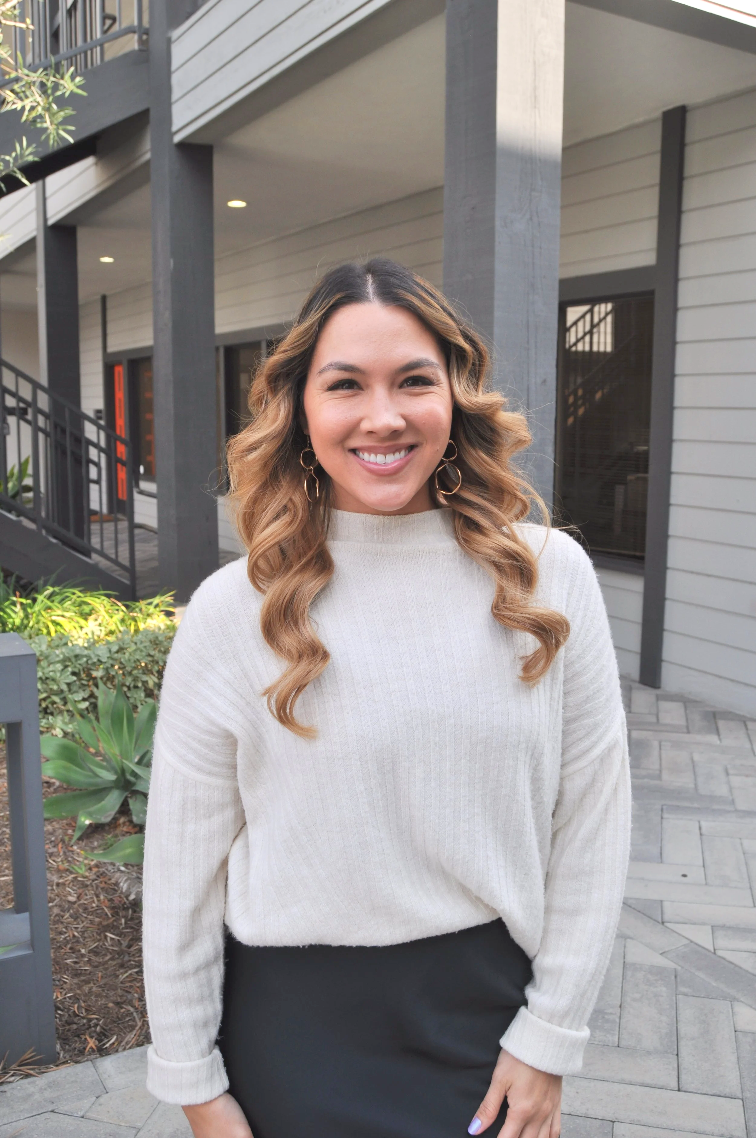 A smiling woman with long, wavy, light brown hair standing outside a modern building with gray siding and black accents.