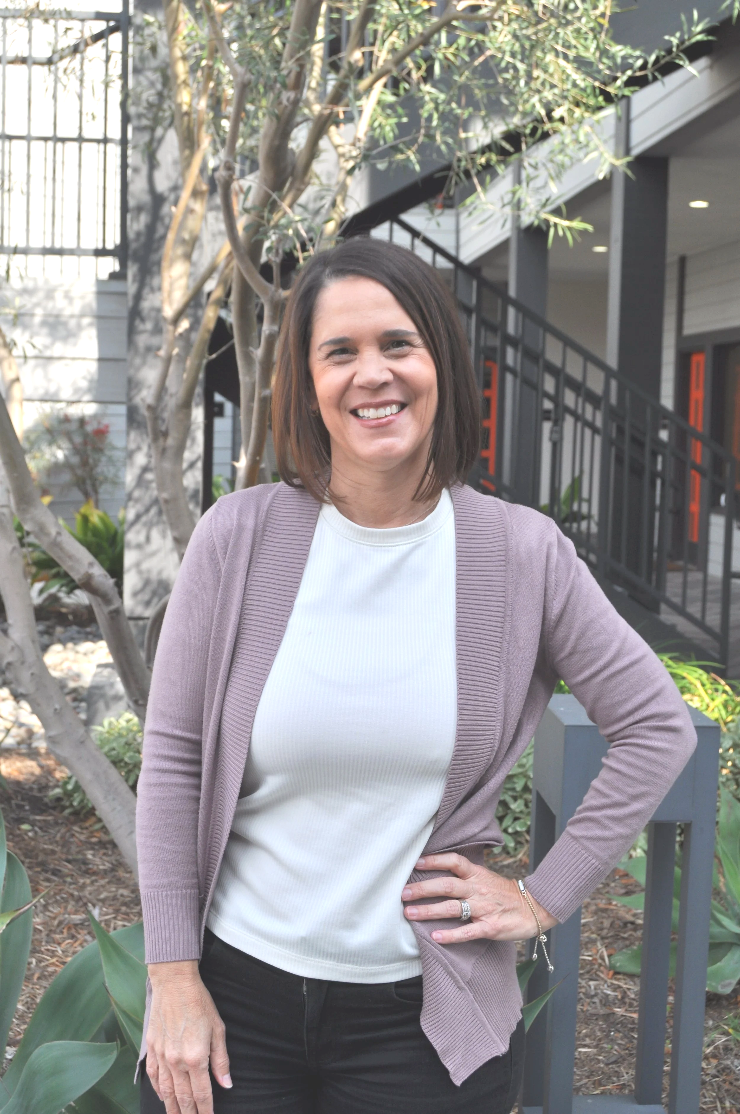 A woman smiling outdoors in front of a tree and a building with a staircase, wearing a white t-shirt and a light purple cardigan.