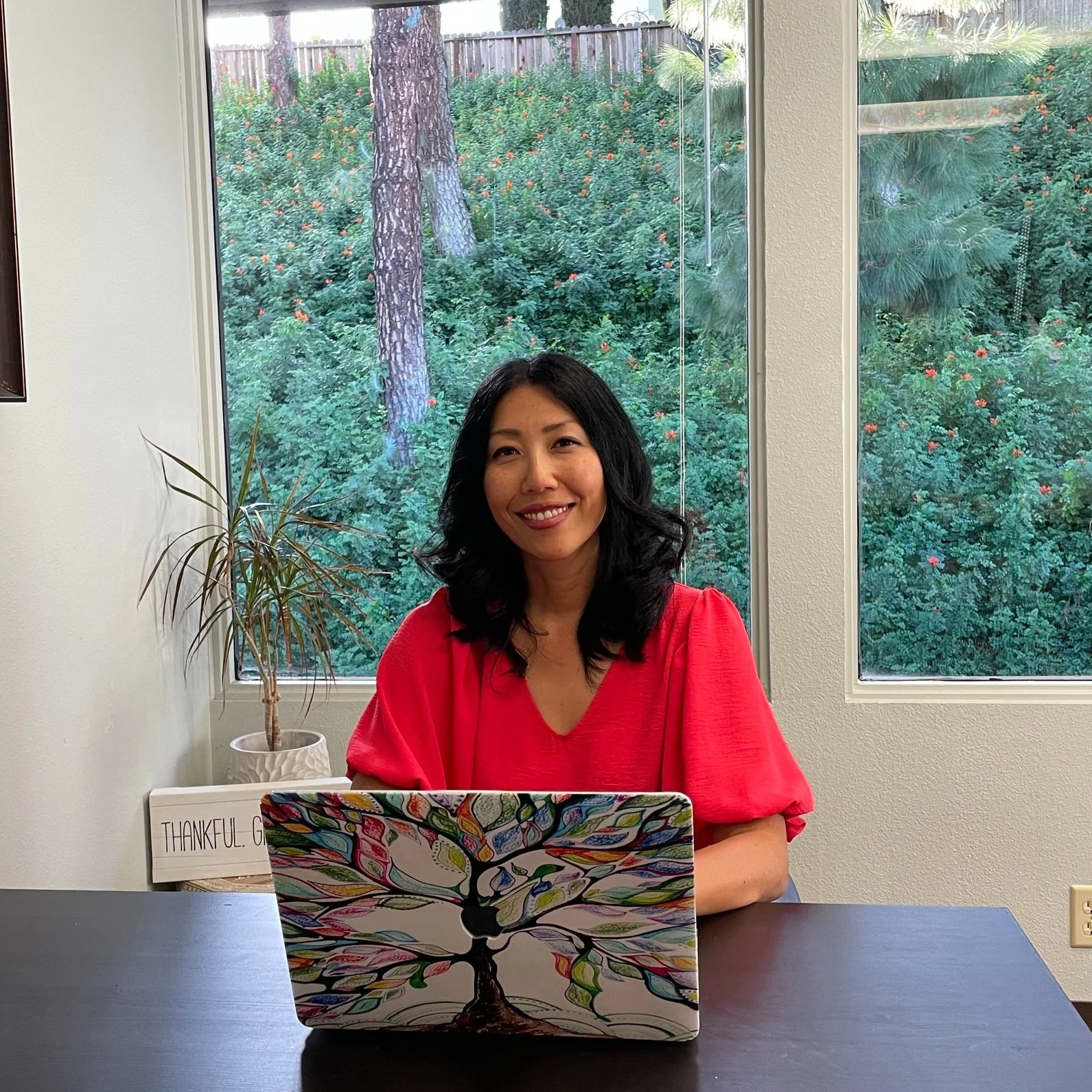 A woman with black hair, wearing a bright pink top, sitting at a table in front of a window with a view of lush greenery and trees outside. There is a colorful tree design on her laptop and a small potted plant beside her. A sign in the background reads 'THANKFUL'.