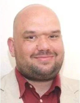 Close-up of a man with a bald head, short beard, wearing a beige blazer and red shirt, smiling at the camera.