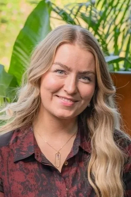 A woman with long, wavy blonde hair smiling, outdoors with green tropical plants in the background.