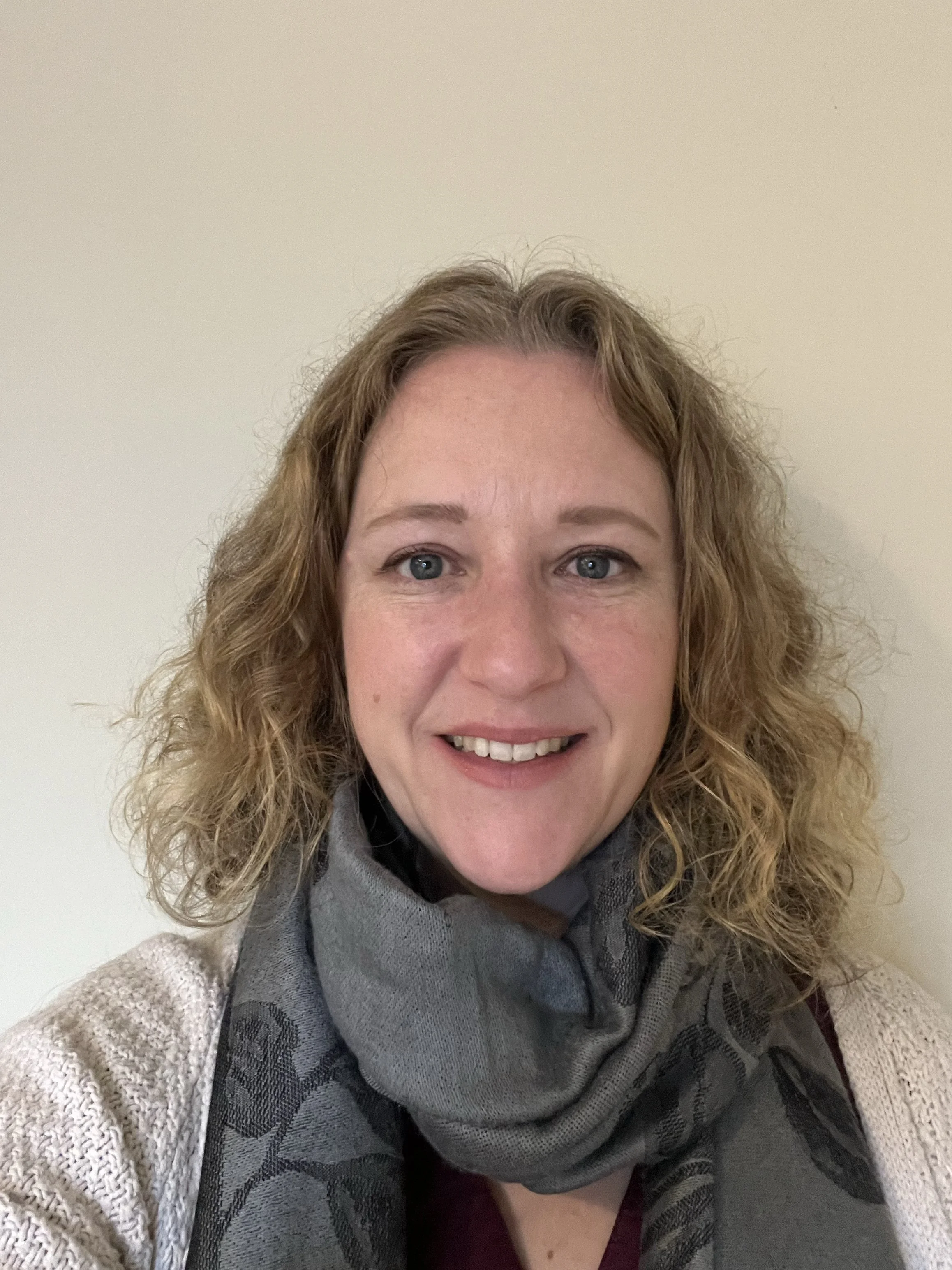 A woman with shoulder-length curly blonde hair smiling, wearing a grey scarf and a cream-colored sweater, standing against a plain beige wall.