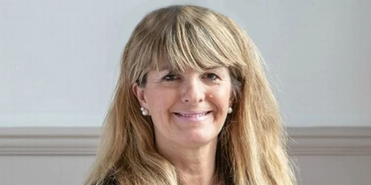 A woman with long blonde hair, wearing pearl earrings, smiling at the camera in front of a light-colored wall.
