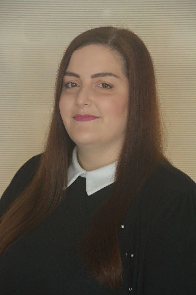 A woman with long brown hair, one eye slightly closed, wearing a black top with a white collar, standing against a light beige horizontal striped background.