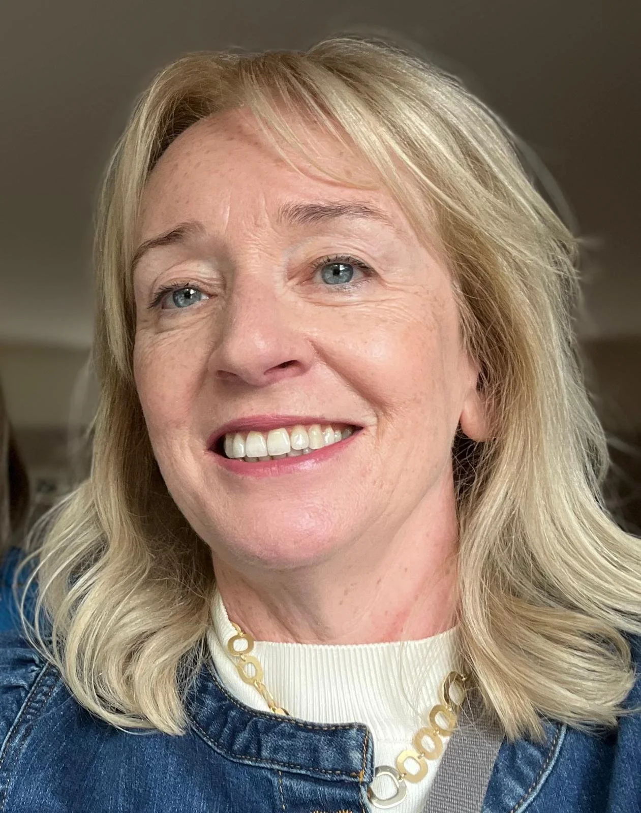 Close-up of a smiling middle-aged woman with blonde hair, blue eyes, wearing a white shirt, gold necklace, and denim jacket.