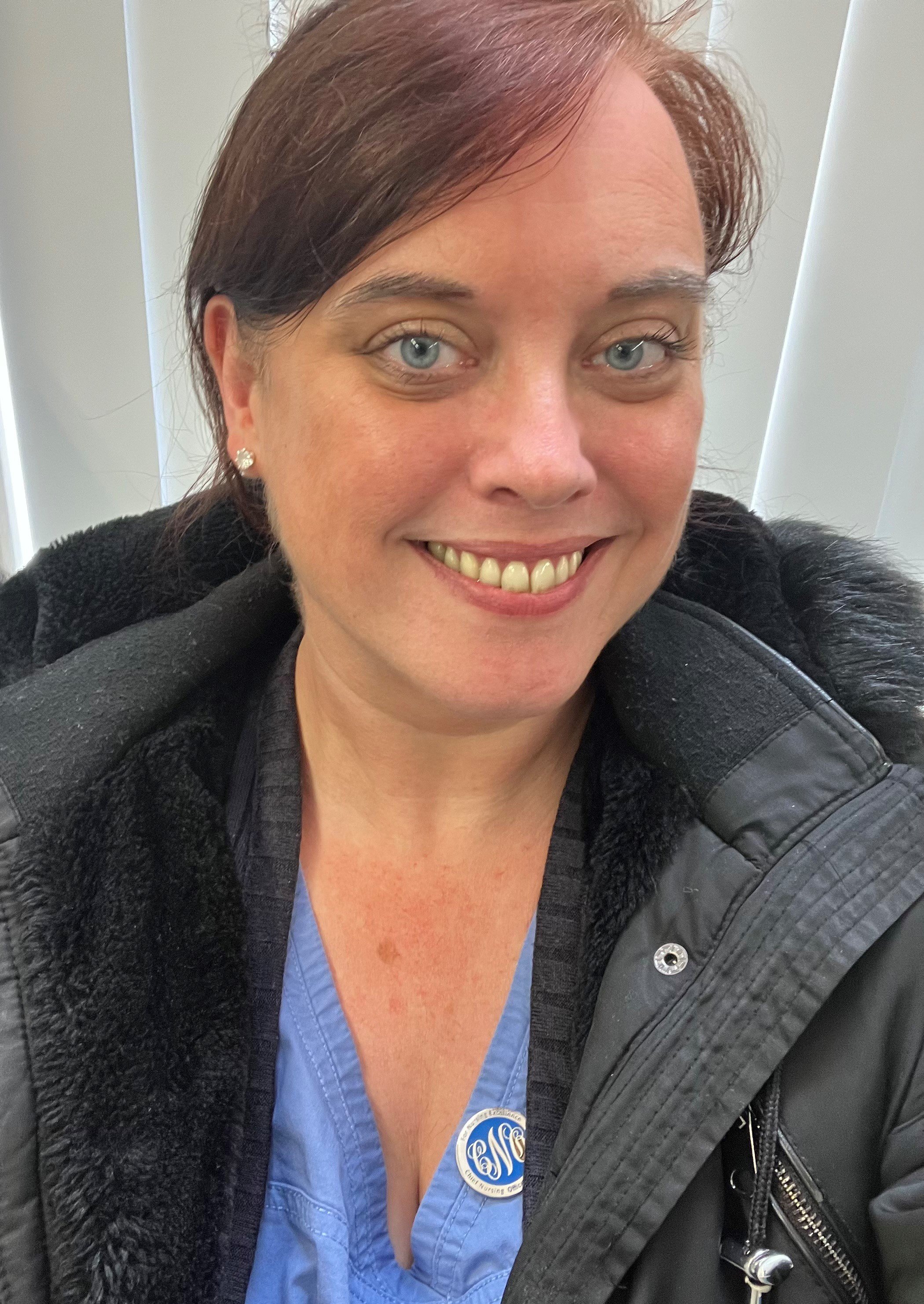 A woman with short dark red hair and blue eyes taking a selfie inside a building, wearing a black jacket with a furry collar and a blue shirt underneath, smiling at the camera.