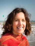 A woman with curly brown hair smiling outdoors near a body of water.