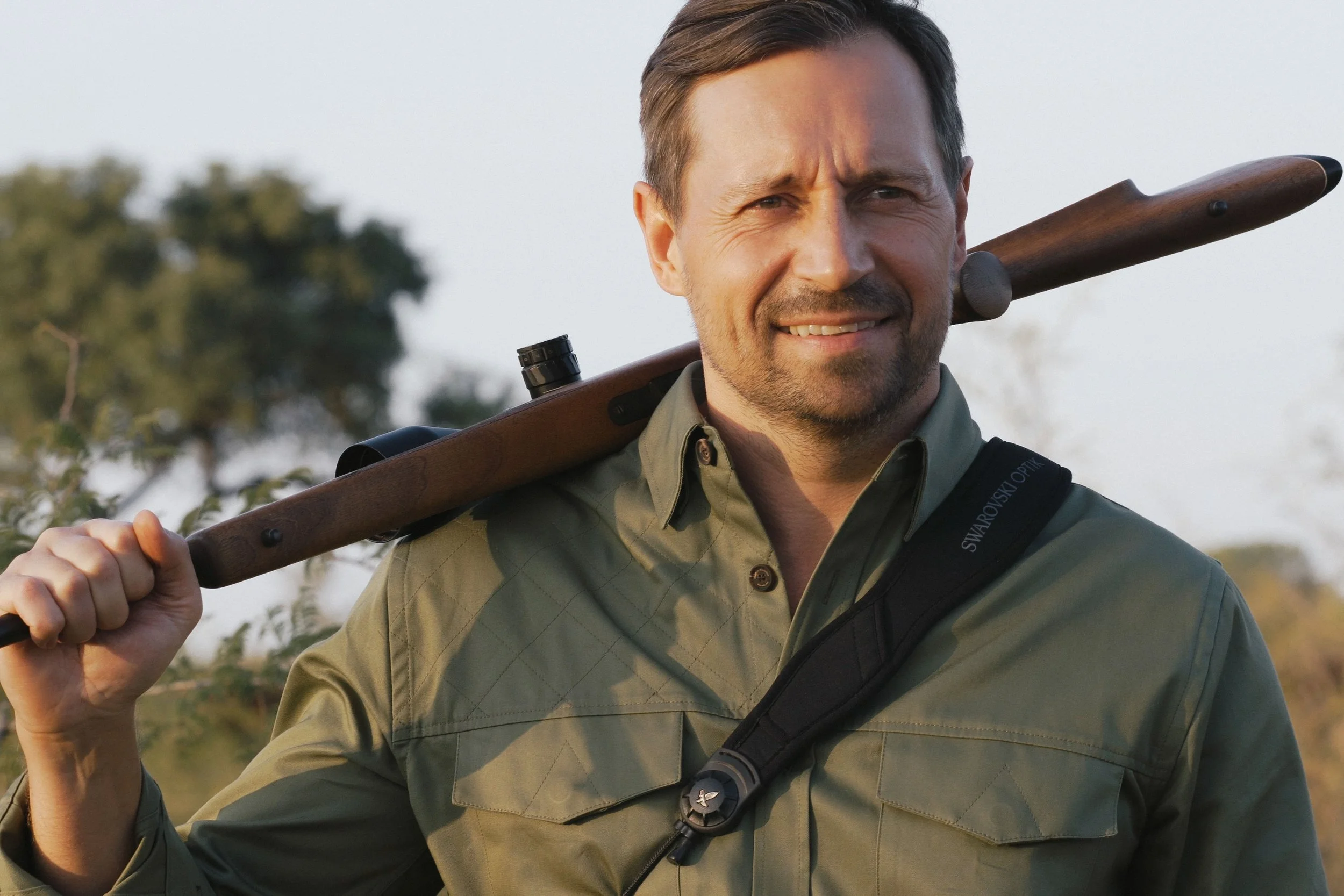 A man outdoors holding a rifle over his shoulder, smiling, wearing a green shirt and a black strap with a Swarovski Optik logo.